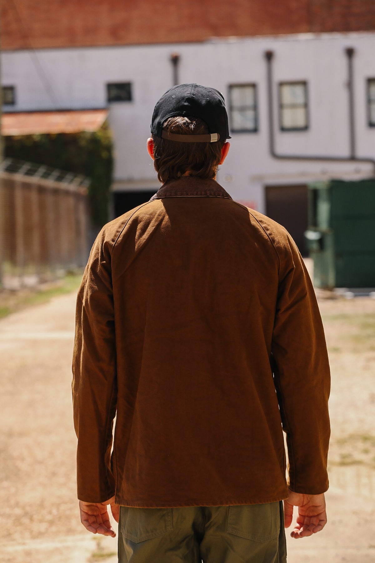 A man viewed from behind wearing a brown canvas utility jacket, a black baseball cap, and olive green pants, standing outdoors on a dirt path with buildings in the background.
