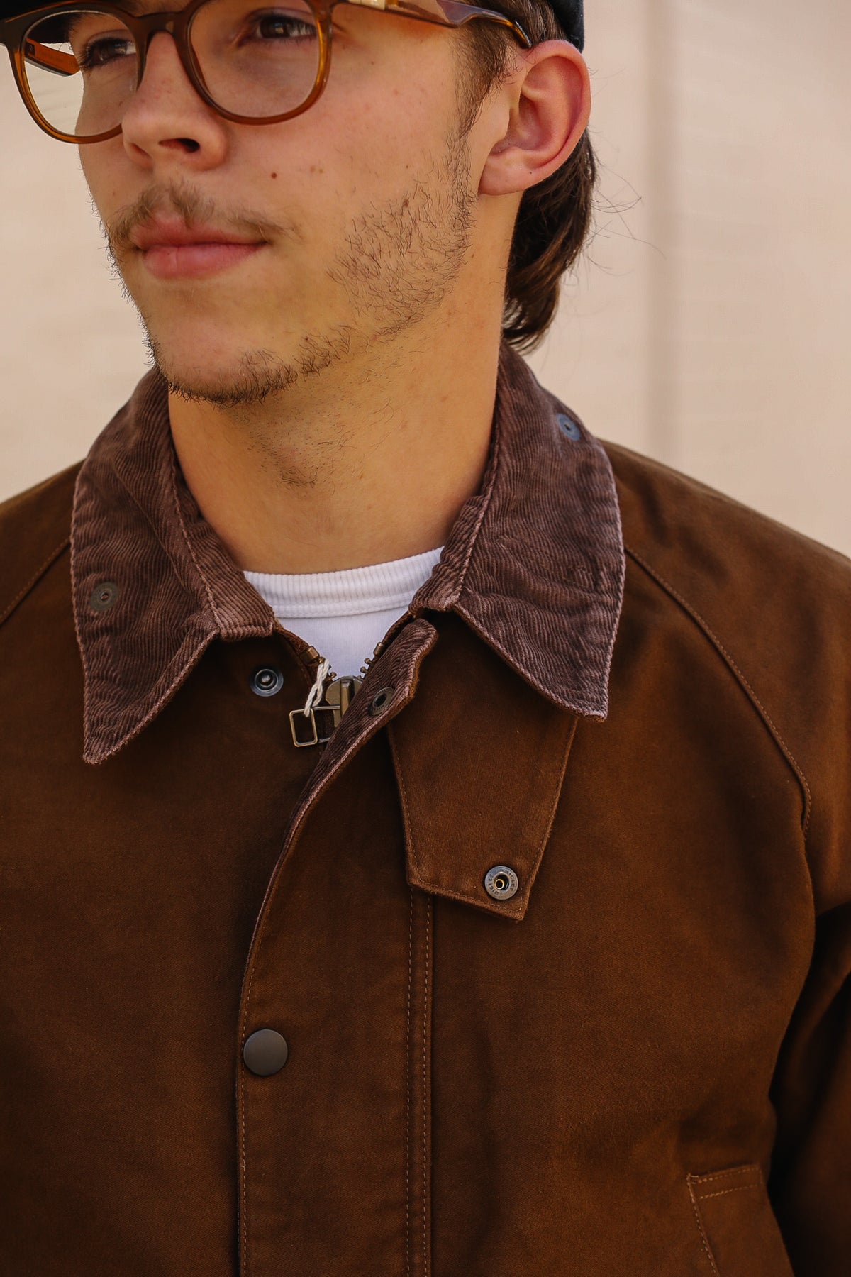 A close-up of a person wearing a brown suede jacket with a contrasting dark brown corduroy collar, layered over a white t-shirt. The person is also wearing glasses and a black cap, set against a neutral beige background.
