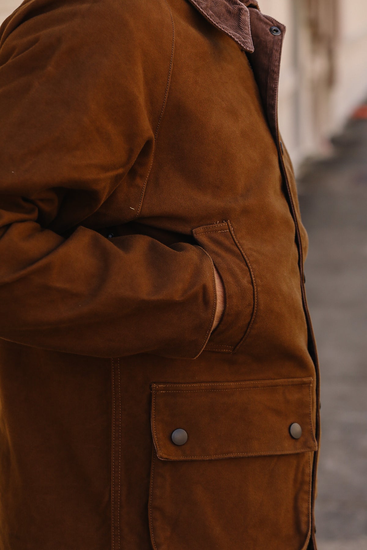Close-up of a person wearing a brown suede jacket with a corduroy collar and flap pockets, photographed outdoors against a blurred street background.