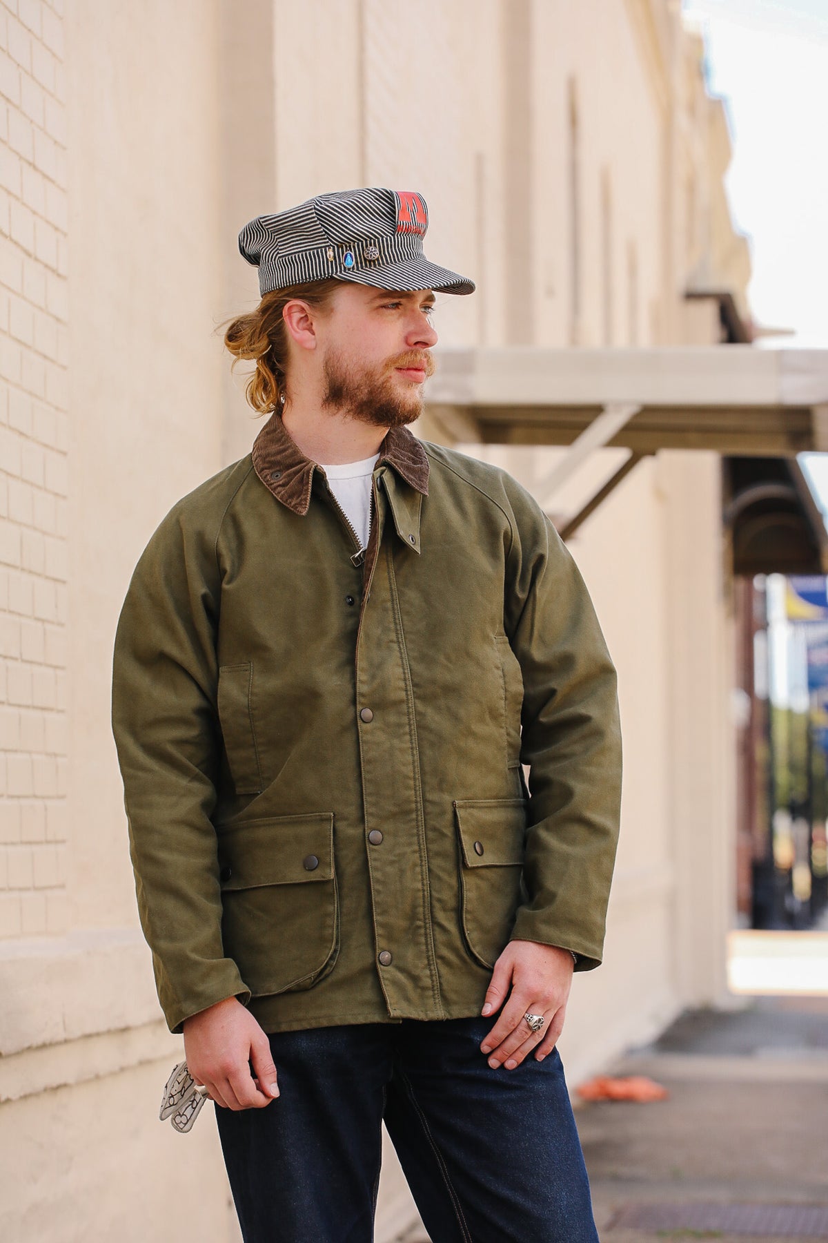 A man wearing an olive green waxed cotton utility jacket with brown corduroy collar, dark blue jeans, and a grey plaid cap, standing outdoors against a beige brick wall.