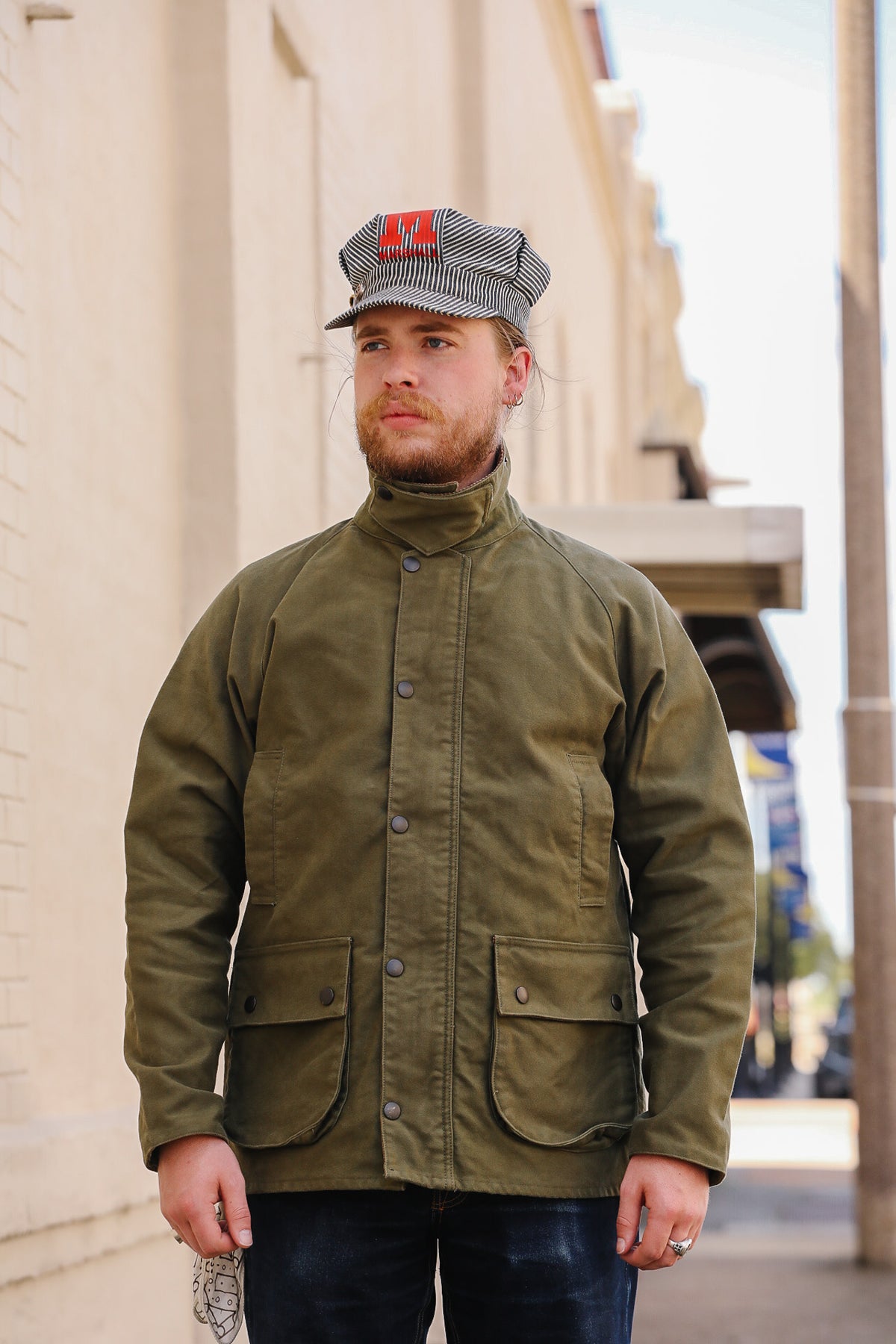 A man wearing an olive green waxed cotton utility jacket and a gray and black checkered cap with a red 'H' logo, standing outdoors against a beige building wall.