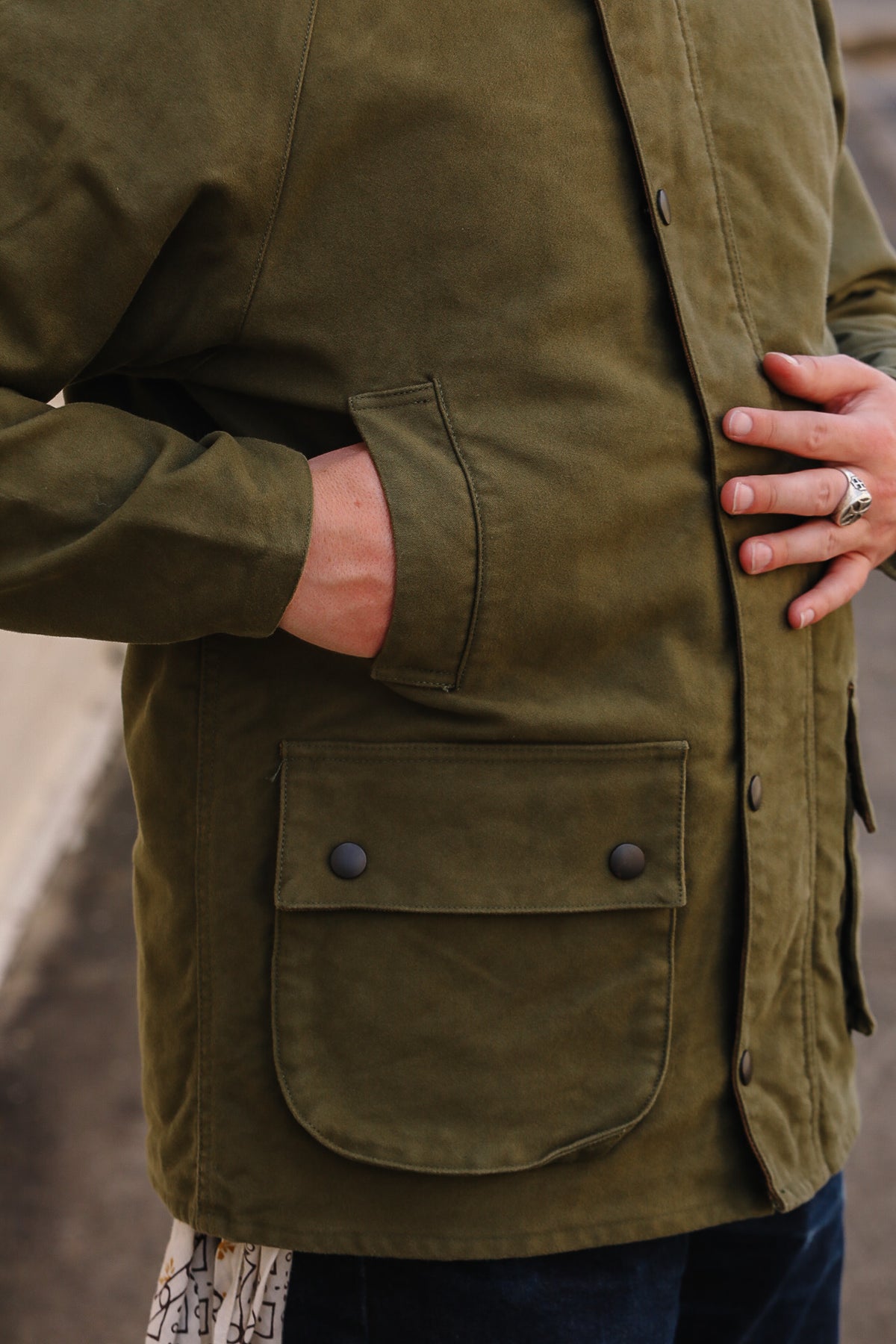 Close-up of a person wearing an olive green waxed cotton jacket, showing a front button closure and a large buttoned patch pocket.