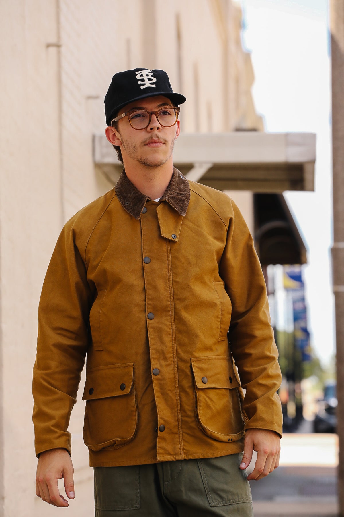 A man wearing a brown waxed cotton field jacket with a dark brown corduroy collar, a black baseball cap, and glasses, standing outdoors on a city street.