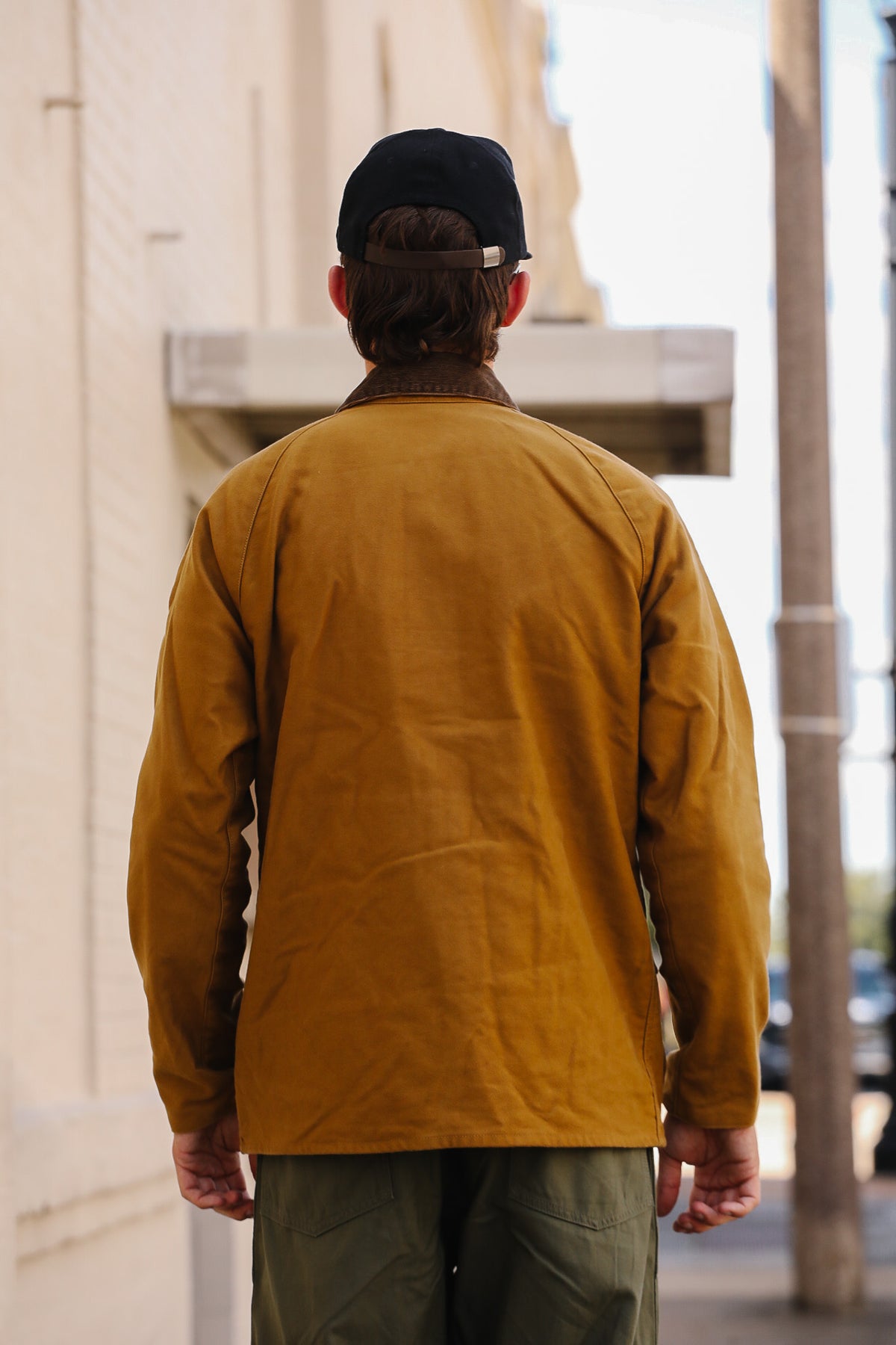 A man viewed from behind wearing a mustard yellow canvas utility jacket, olive green pants, and a black baseball cap on a city sidewalk.