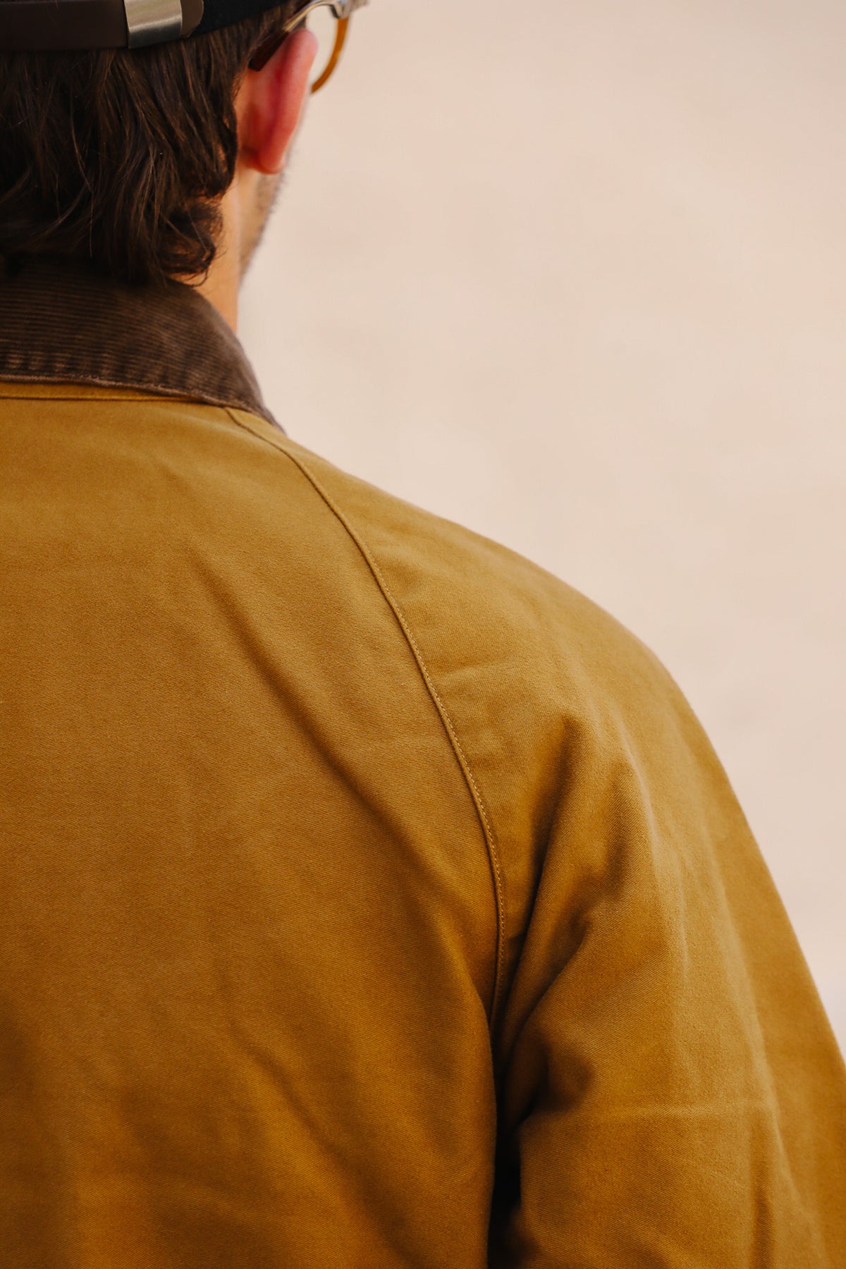 Close-up back view of a person wearing a mustard yellow jacket with a dark brown collar, photographed against a plain light beige background.
