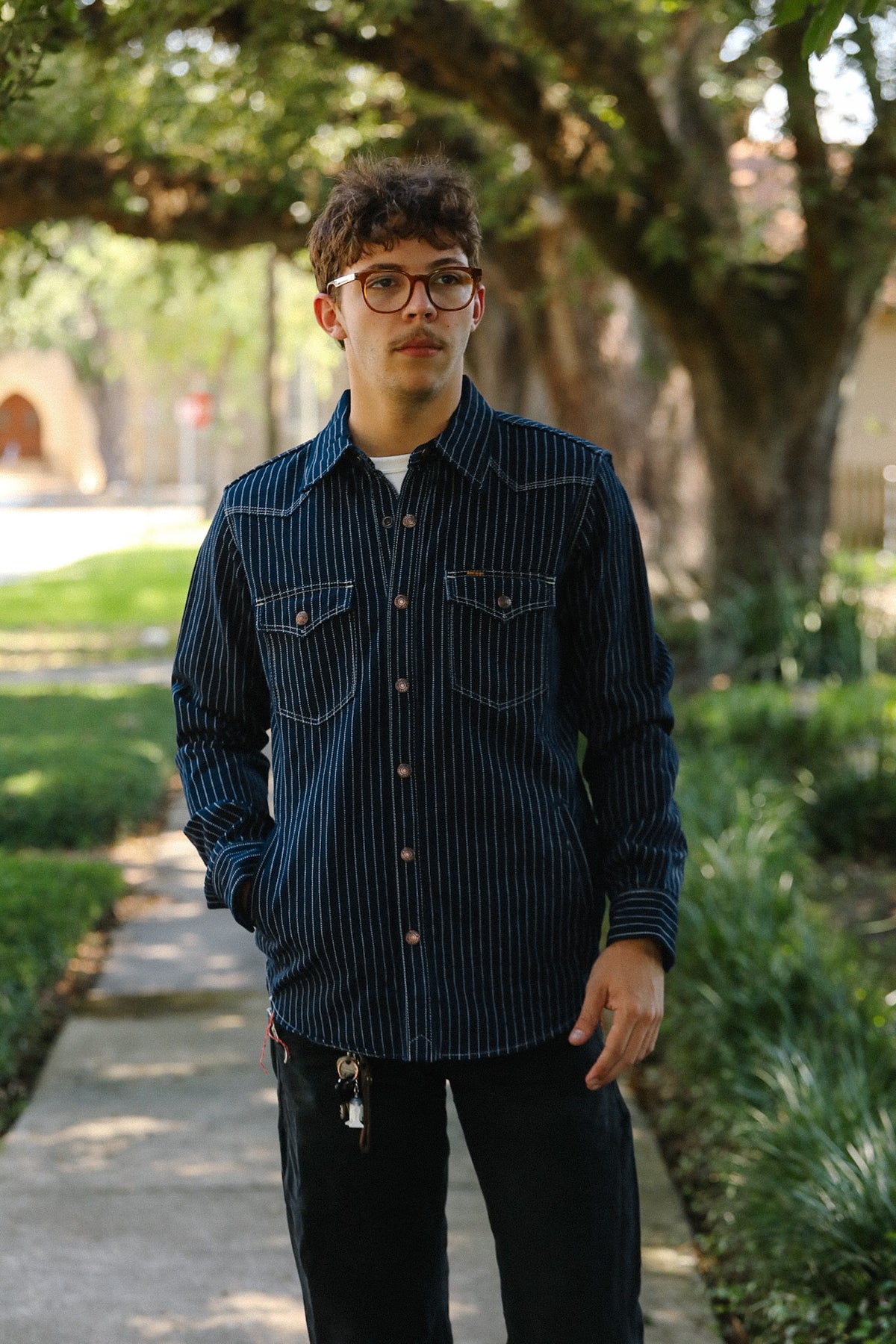 A man wearing a dark navy blue pinstriped button-down shirt and dark jeans, standing outdoors on a paved path surrounded by green trees and grass.