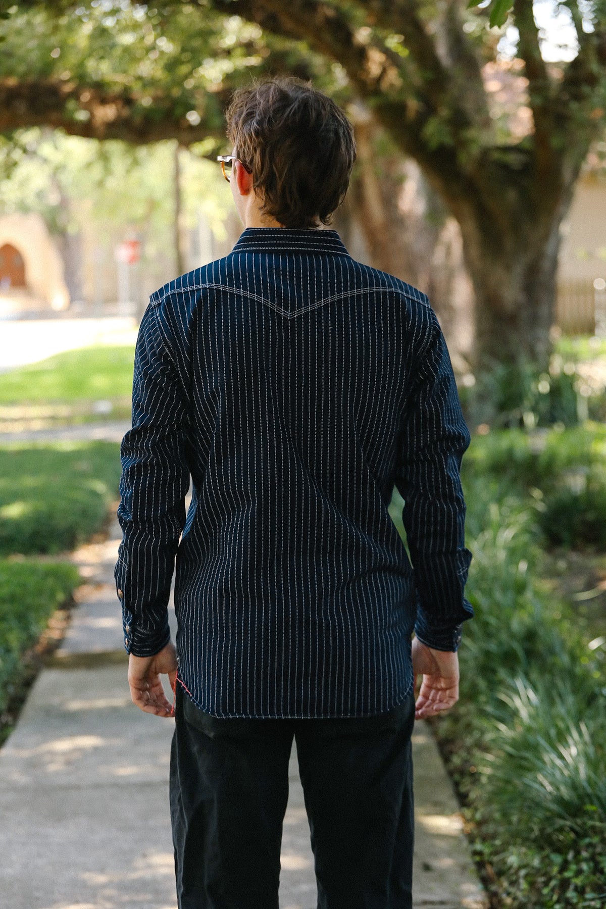 A man viewed from behind wearing a dark navy blue long-sleeve shirt with thin vertical white pinstripes and black pants, standing on a paved path in a garden setting with trees and greenery in the background.