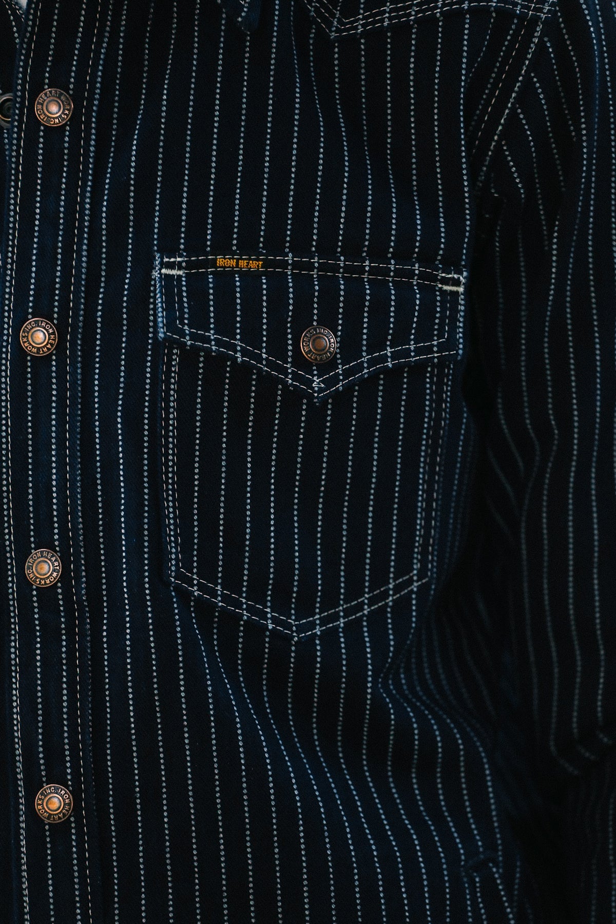 Close-up of a dark navy pinstriped shirt with white stitching, featuring a chest pocket with a button and decorative brass-tone buttons down the front.