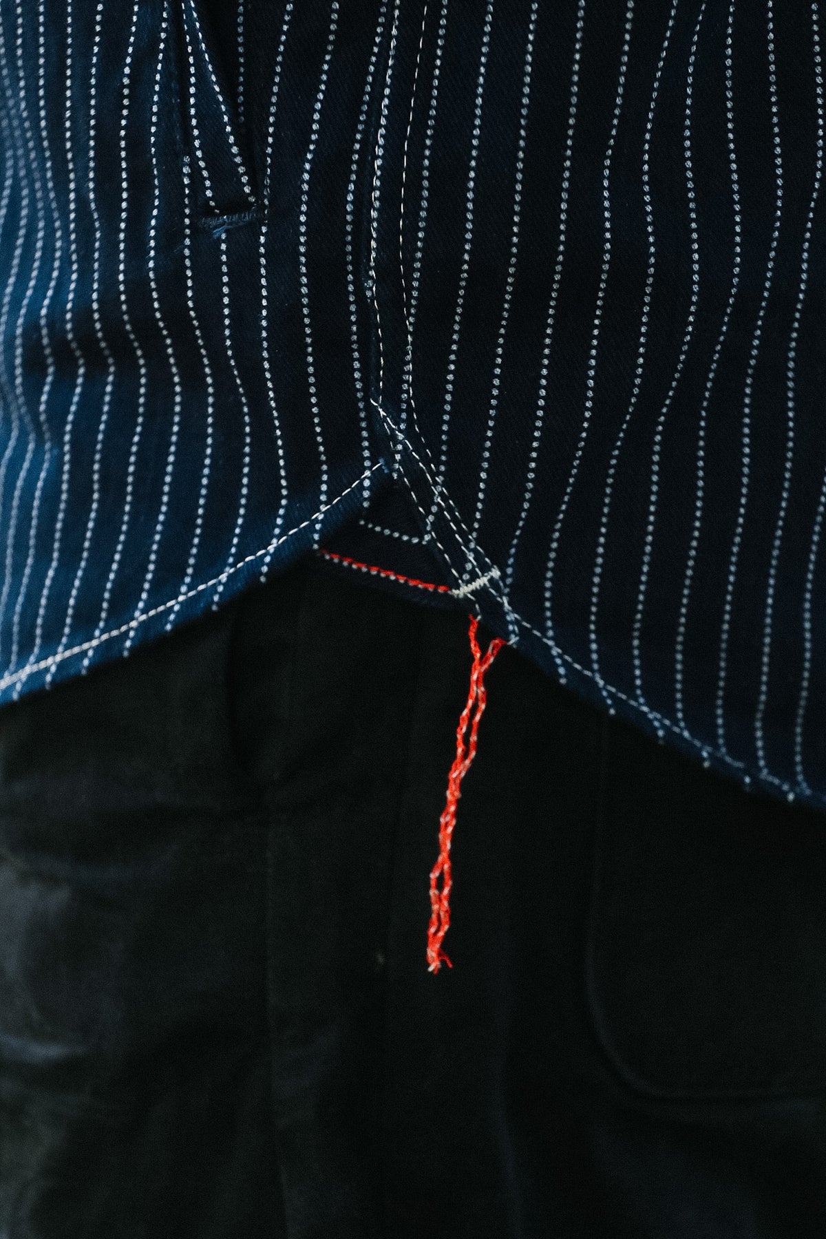 Close-up of a dark navy pinstriped jacket with white contrast stitching and a bright orange-red thread tail hanging from the hem, paired with black trousers.