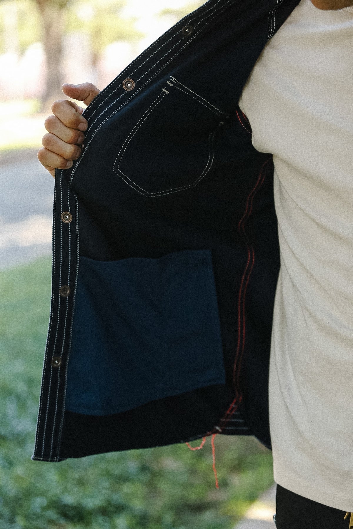 Close-up of a person wearing a dark indigo denim jacket, holding the collar open to show the interior lining, contrast stitching, and a blue patch pocket, with a white t-shirt visible underneath.