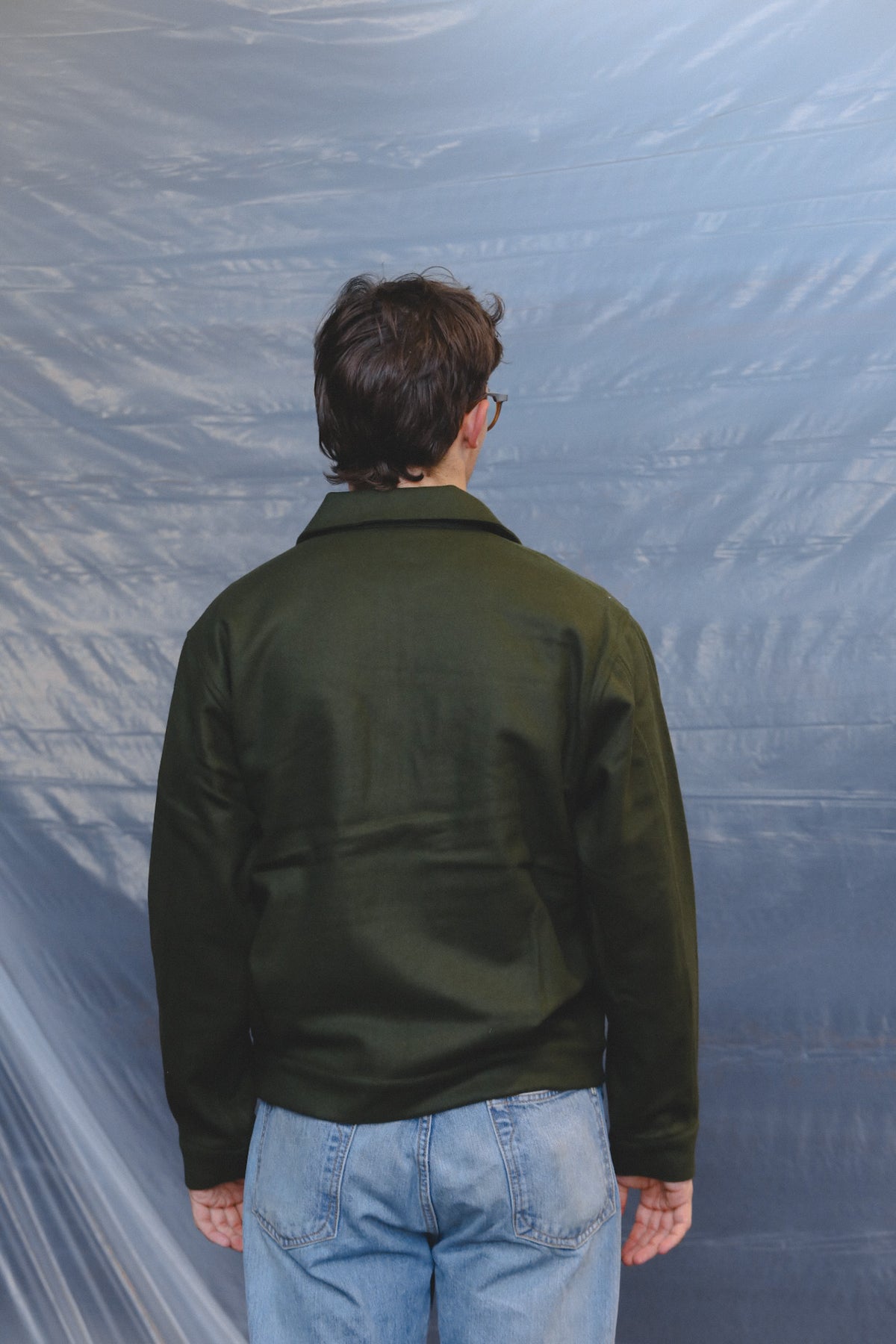 A man viewed from behind wearing an olive green jacket and light blue jeans, standing against a wrinkled blue-gray backdrop.