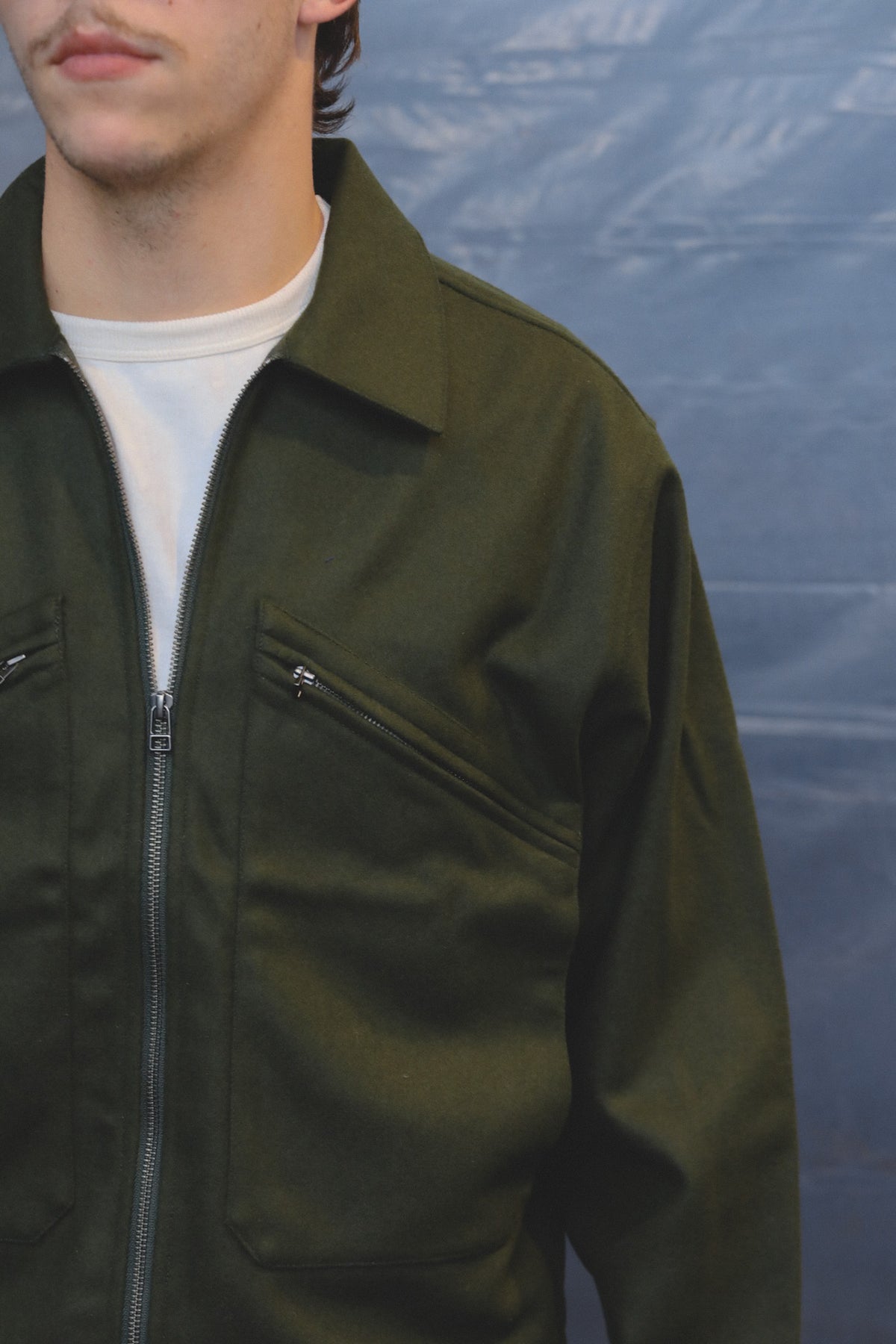 A close-up of a person wearing an olive green zip-front jacket over a white t-shirt, set against a textured blue backdrop.