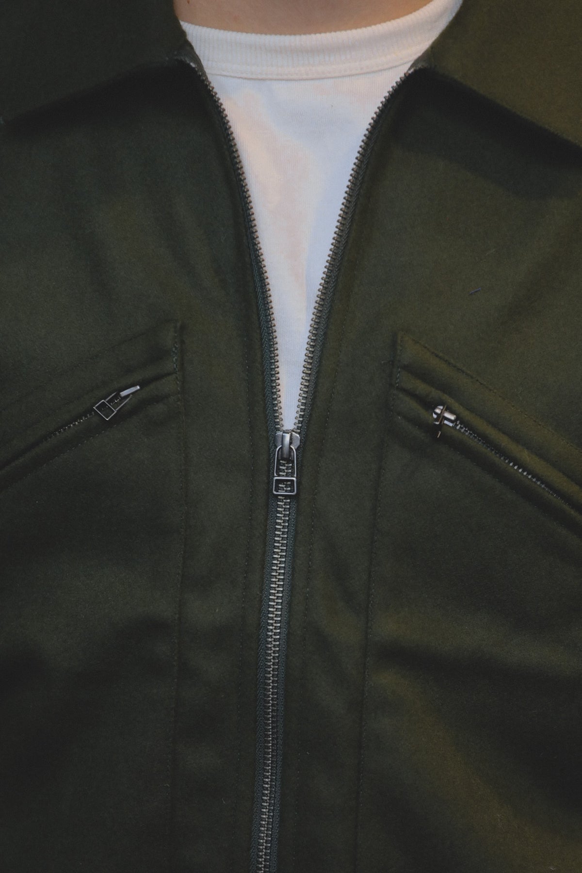 Close-up of a dark olive green zip-up jacket worn over a white t-shirt, showing the front zipper, zippered chest pockets, and collar.