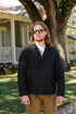 Man wearing black button-up jacket and sunglasses standing outdoors with house and tree in background
