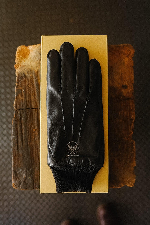 A black leather glove with a ribbed knit cuff, displayed on top of a gold-colored rectangular box. The box is placed on two rustic wooden blocks over a dark textured floor.