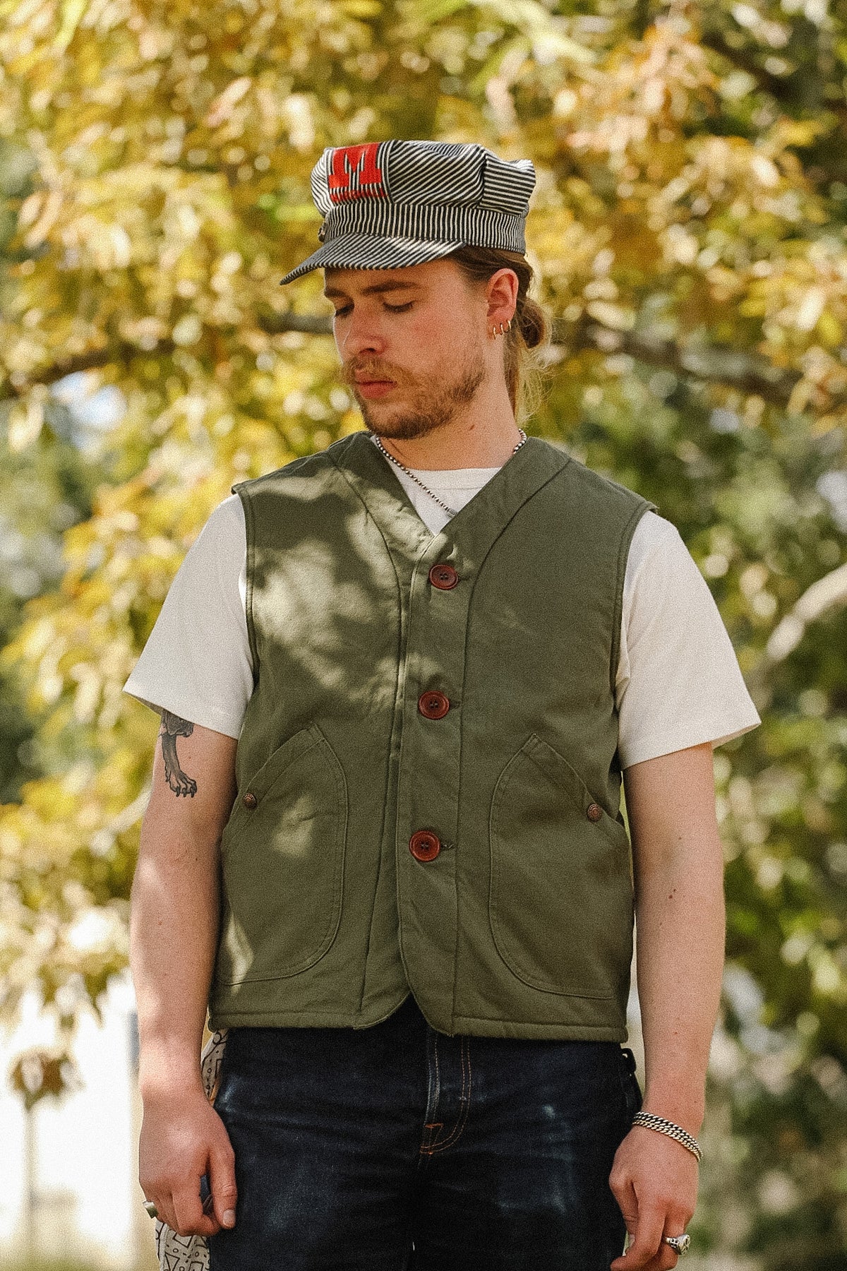 A man wearing an olive green button-up vest over a white t-shirt, dark jeans, and a gray patterned cap with red lettering, standing outdoors in front of a blurred background of green and yellow autumn trees.