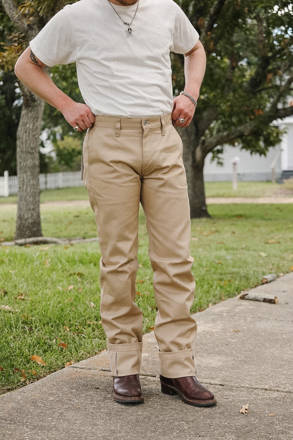 A person standing outdoors on a concrete path wearing beige cuffed pants, a white t-shirt, and brown boots, with grass and trees in the background.