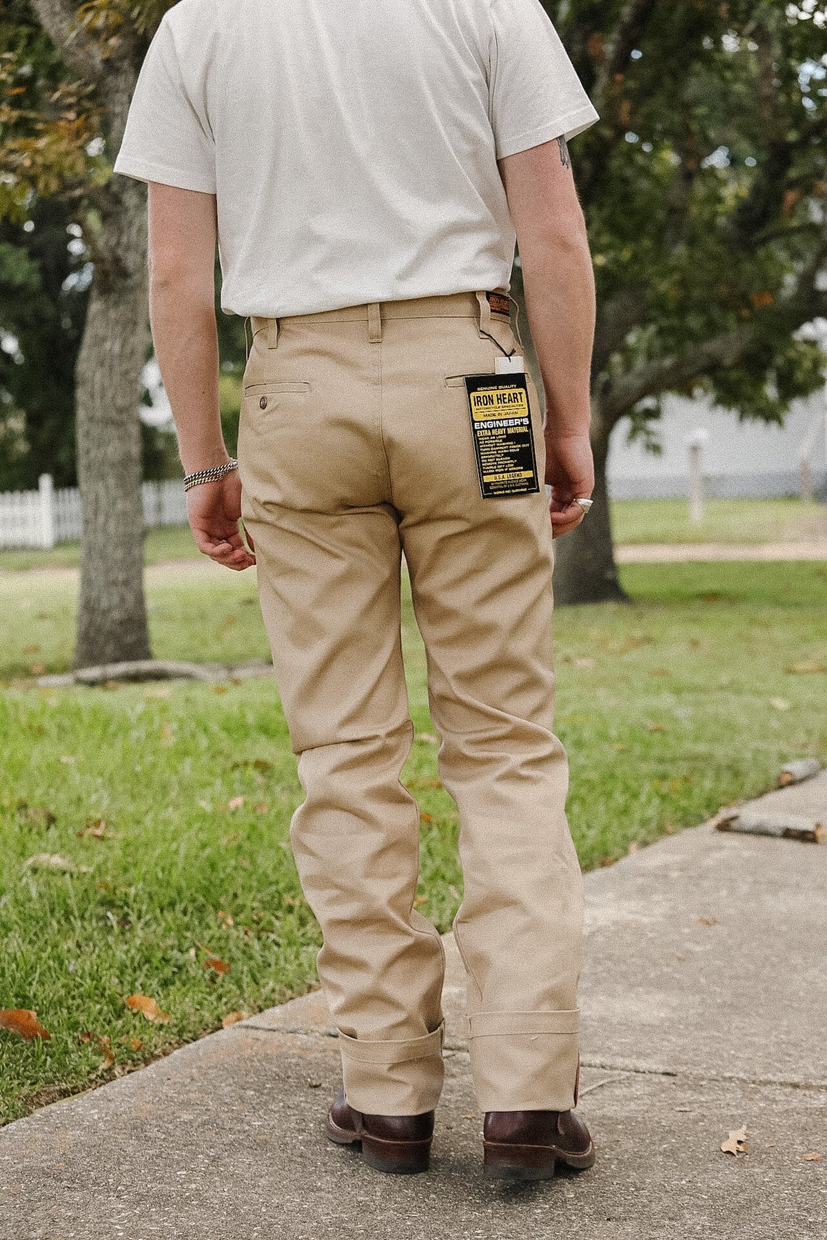 A man viewed from behind wearing khaki work pants, a white t-shirt, and brown shoes, standing on a concrete path in a grassy park area. A yellow and black tag is visible on the back pocket of the pants.