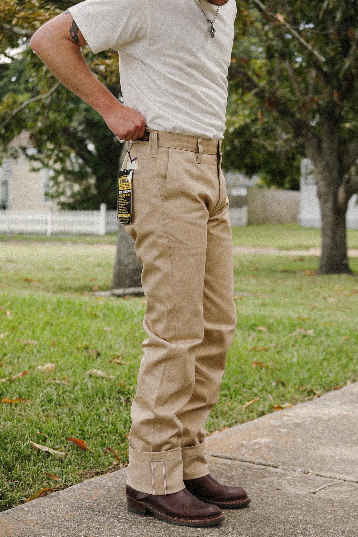 A person outdoors wearing khaki work pants, a white t-shirt, and dark brown boots, standing on a concrete sidewalk with grass and trees in the background.