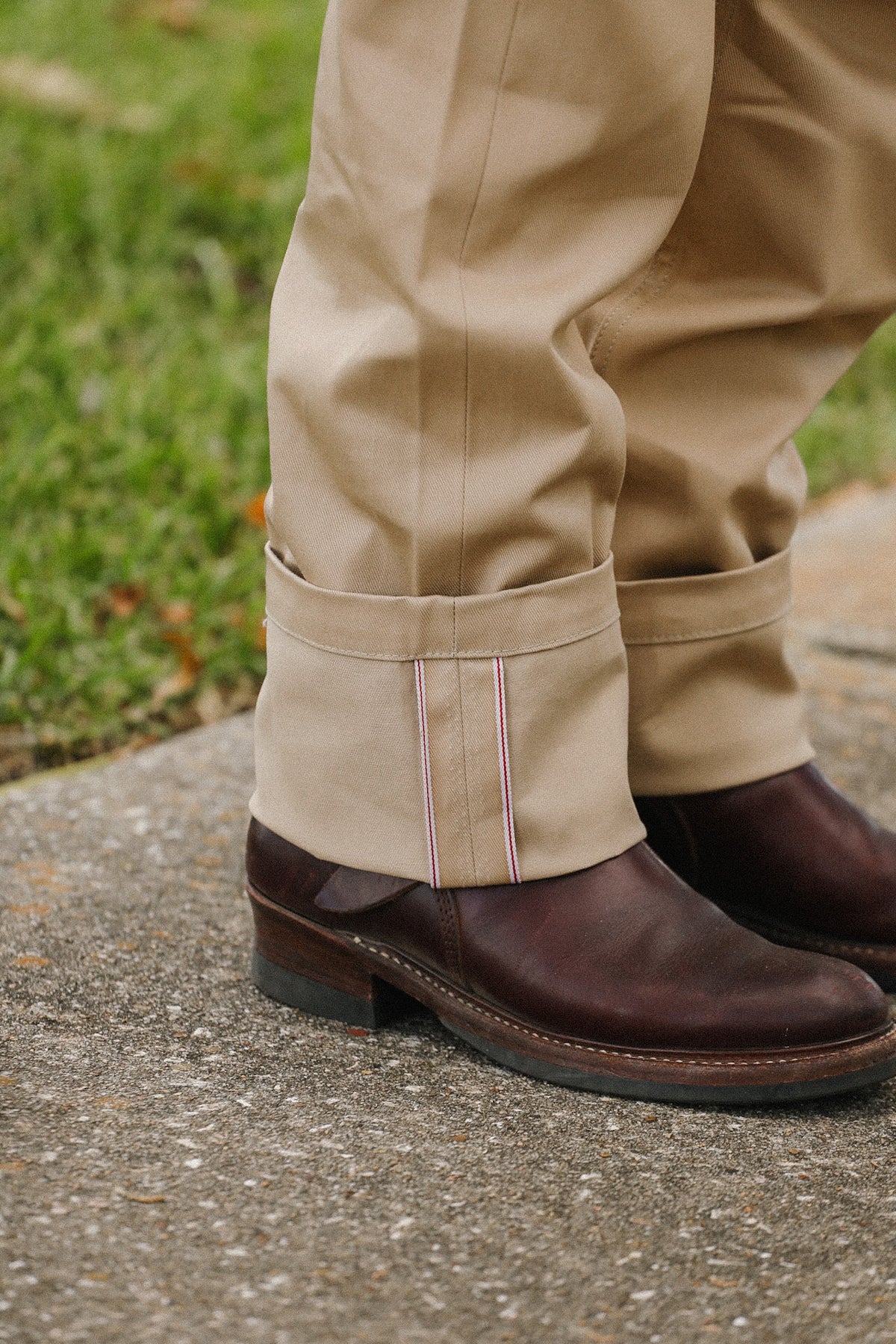 Close-up of a person's legs wearing beige rolled-up trousers with a thin red and white stripe detail, paired with dark brown leather boots on a concrete surface with grass in the background.