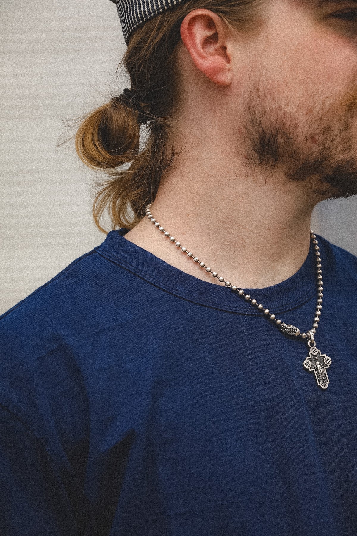 A close-up side profile of a man with a beard and a small ponytail, wearing a silver beaded necklace with a cross pendant over a dark blue t-shirt.