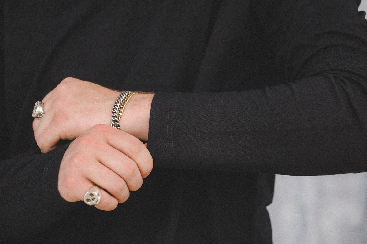 A close-up of a person's hands wearing multiple jewelry pieces, including a silver chain bracelet, a silver ring with a skull design, and another silver ring, all worn over a black long-sleeve shirt.