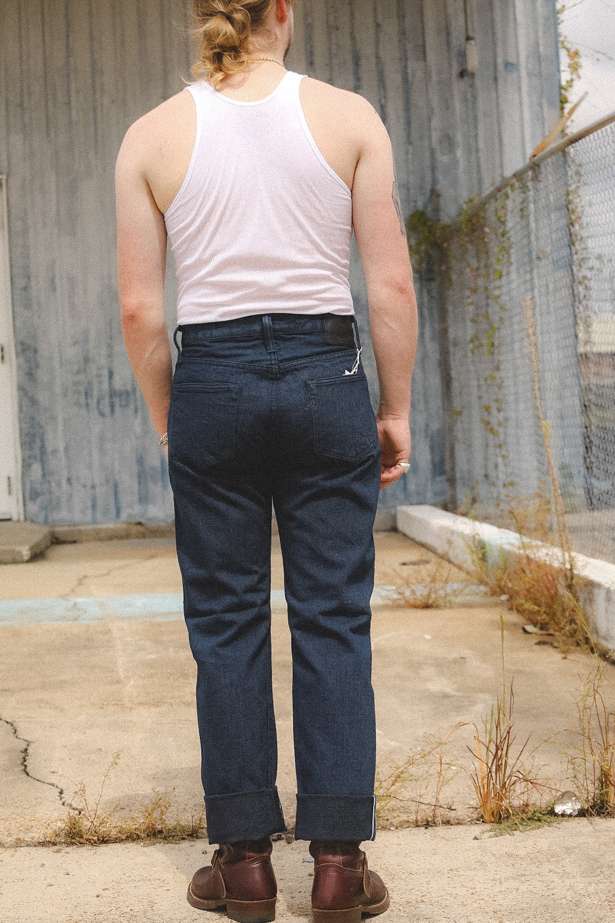 A person standing outdoors with their back to the camera, wearing a white ribbed tank top, dark blue cuffed denim jeans, and brown boots in front of a weathered gray metal wall.