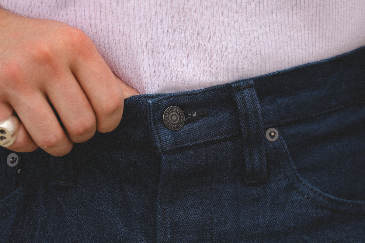 Close-up of a person's hand pulling the waistband of dark blue denim jeans, showing a light pink ribbed knit top underneath.