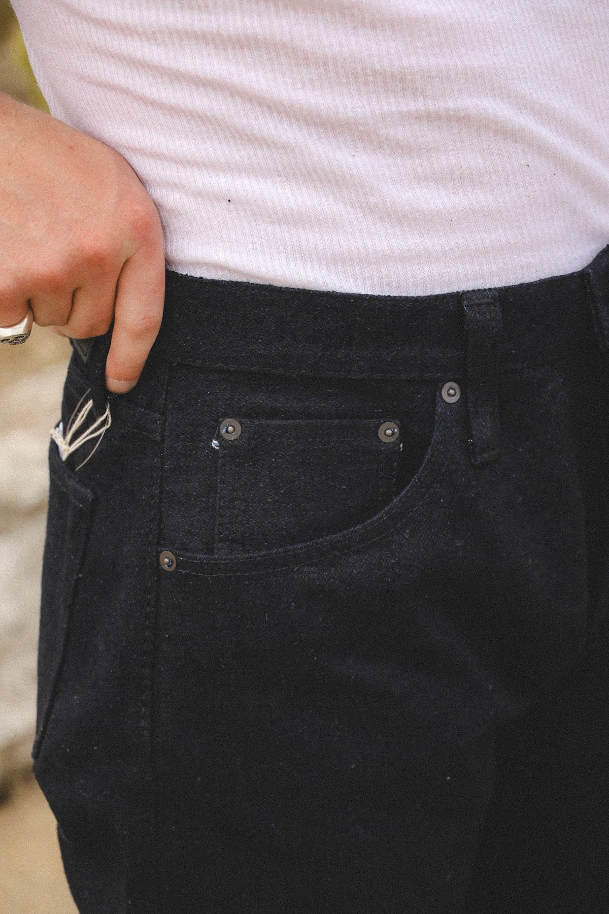 Close-up of a person wearing dark indigo denim jeans with a white ribbed t-shirt, showing the waistband, pocket details, and a hand pulling the fabric.