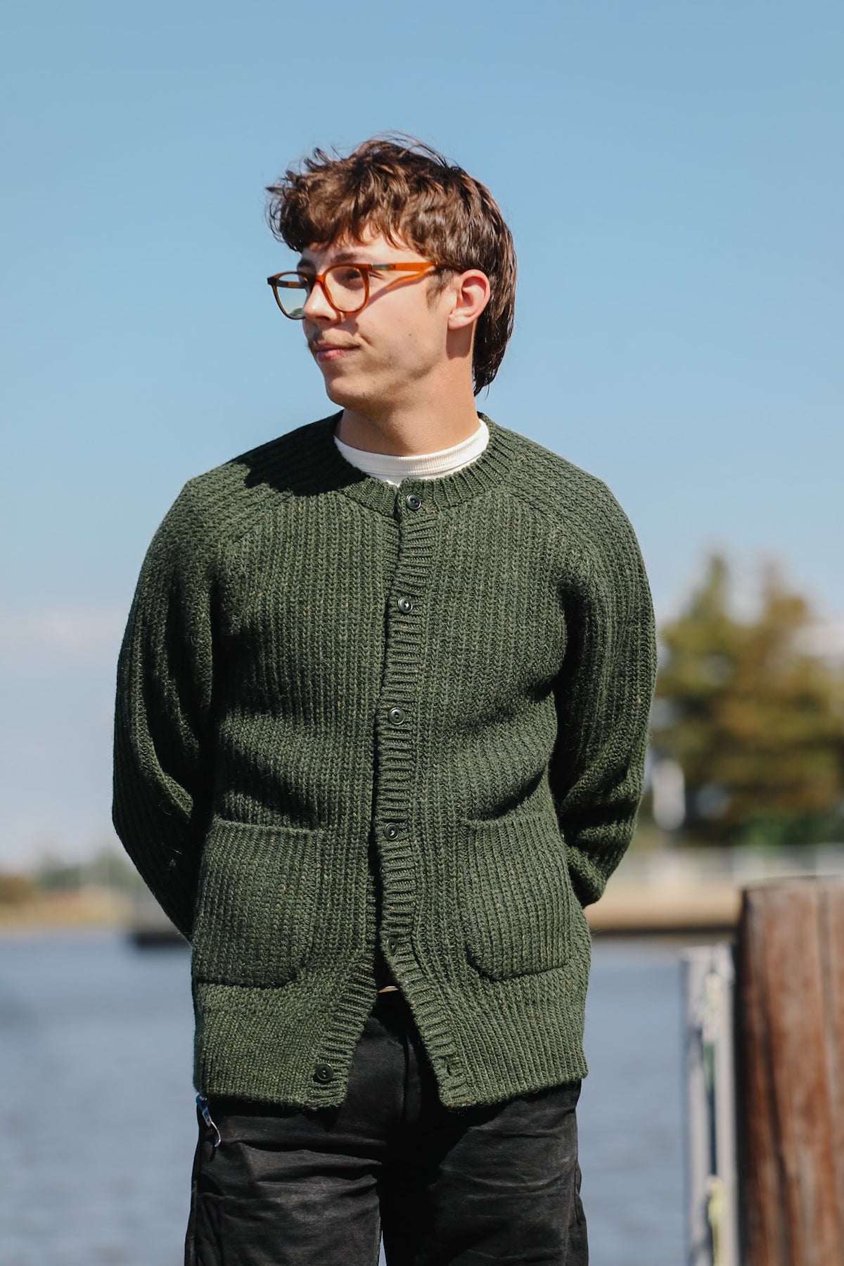 A man wearing a dark green ribbed knit cardigan sweater with front pockets, standing outdoors near water under a clear blue sky.
