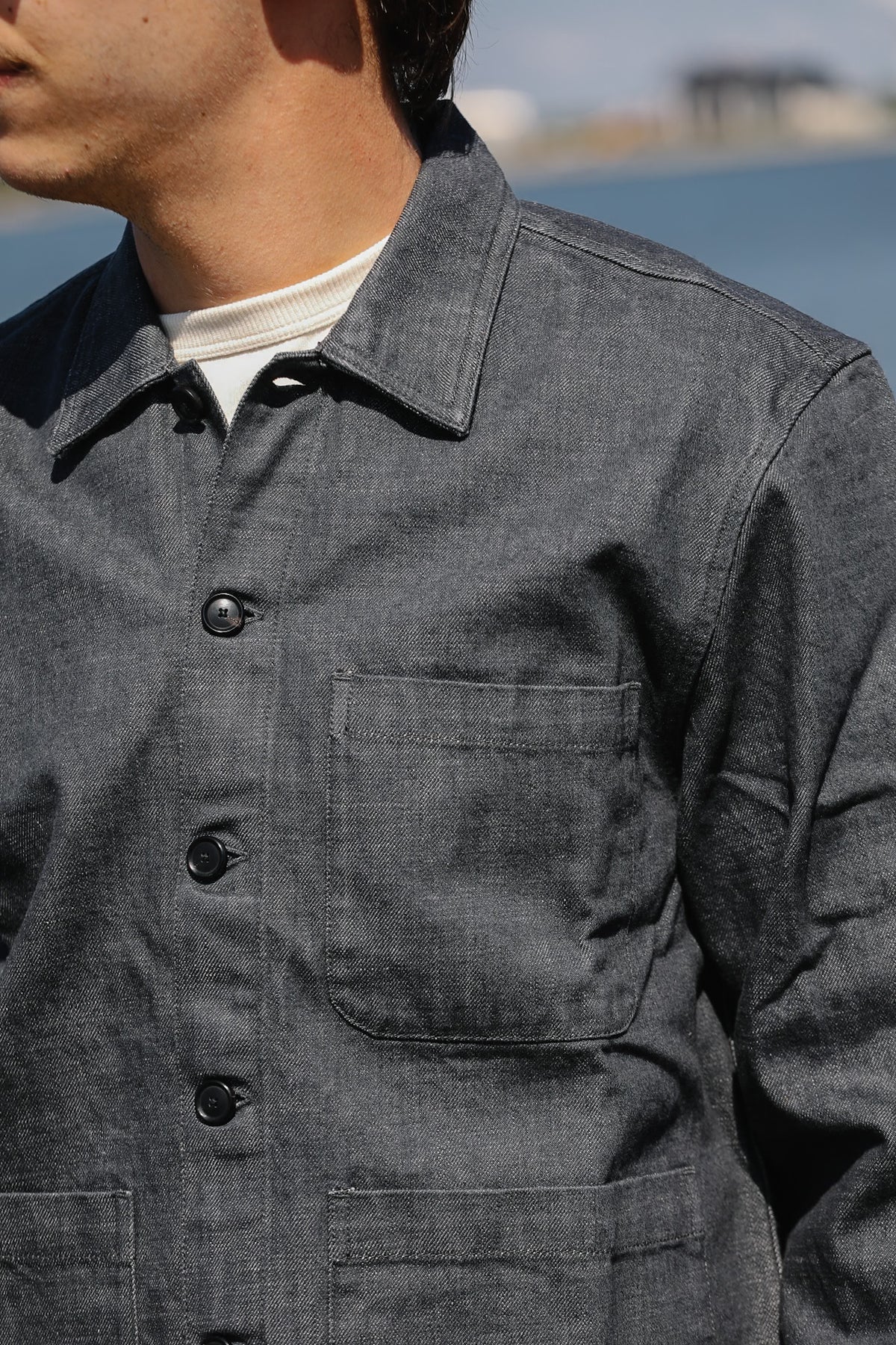 A close-up of a person wearing a dark gray textured overshirt or jacket over a white t-shirt, with a blurred outdoor background.