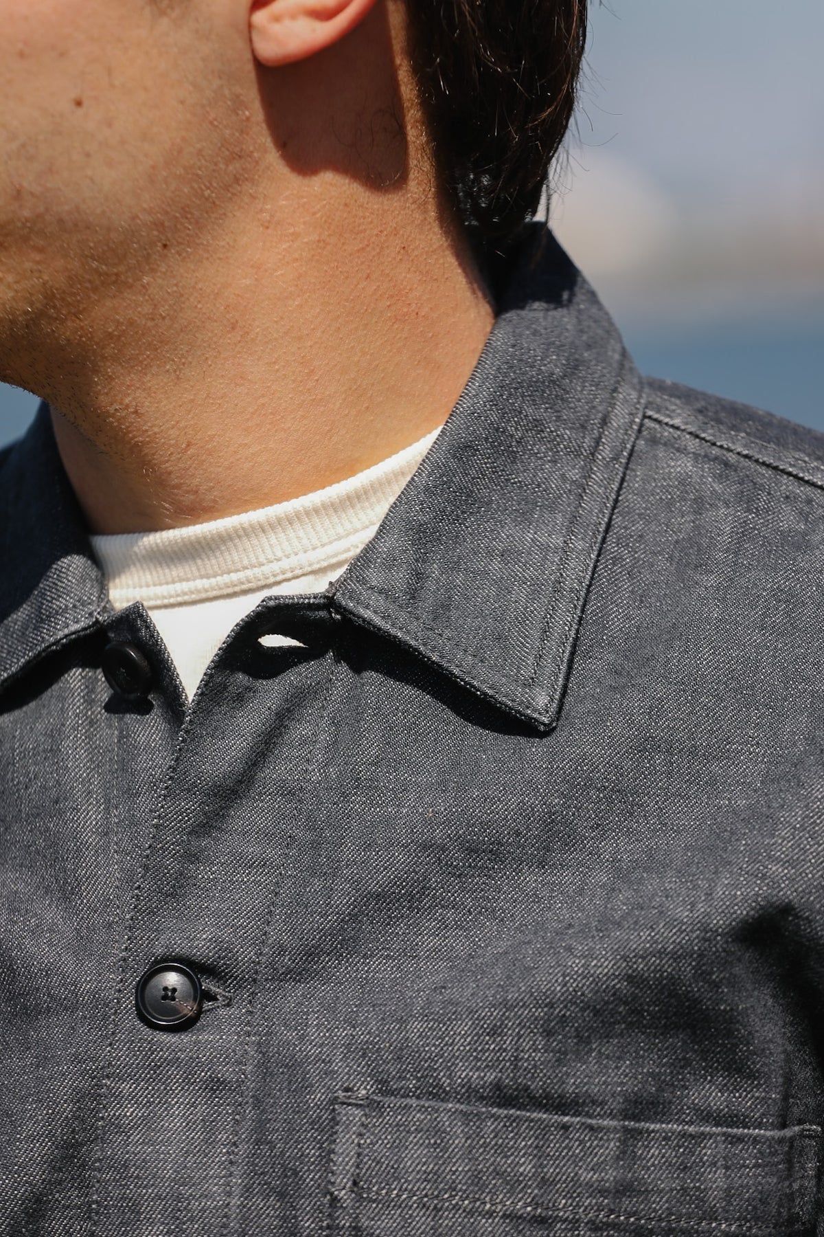 Close-up of a person wearing a dark gray textured overshirt over a white crewneck shirt, with a blurred outdoor background.