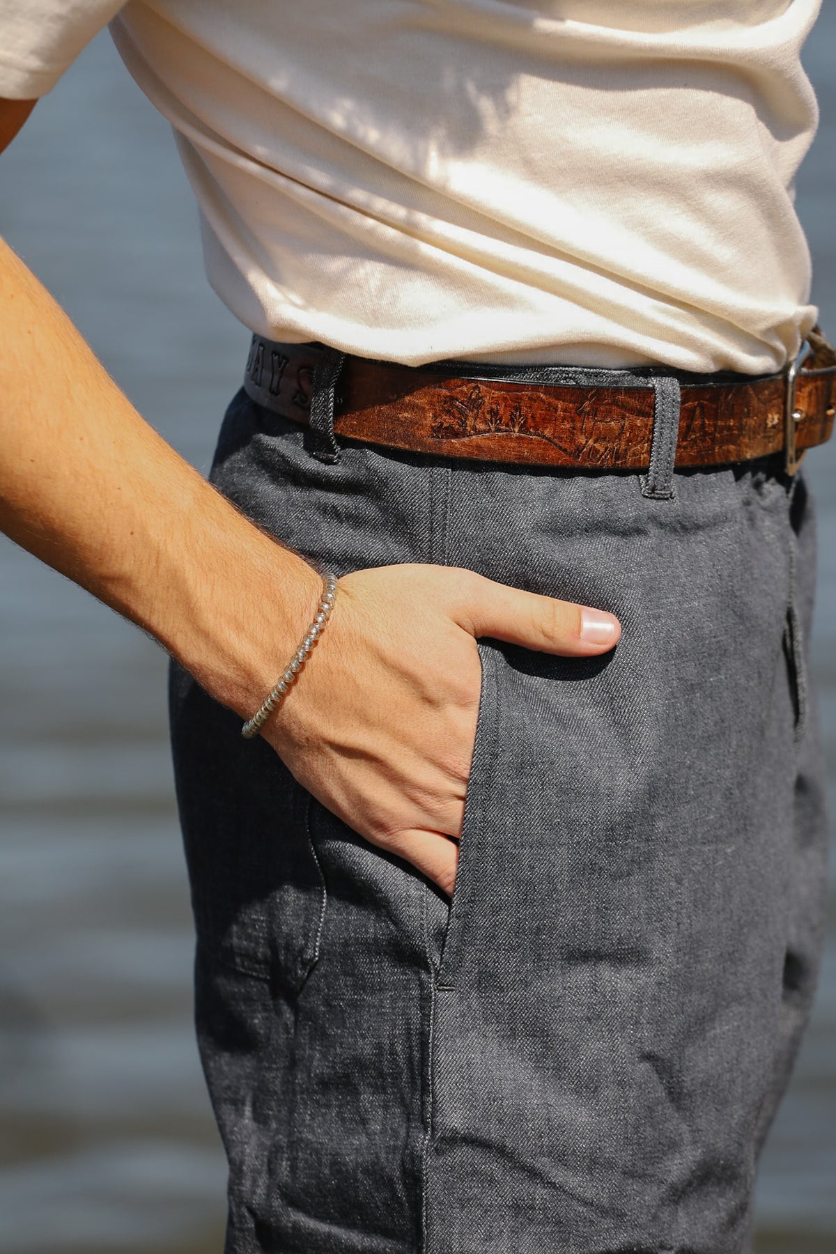 Close-up of a person wearing dark gray linen trousers, a brown patterned leather belt, and a cream-colored t-shirt, with one hand in the pocket of the trousers.