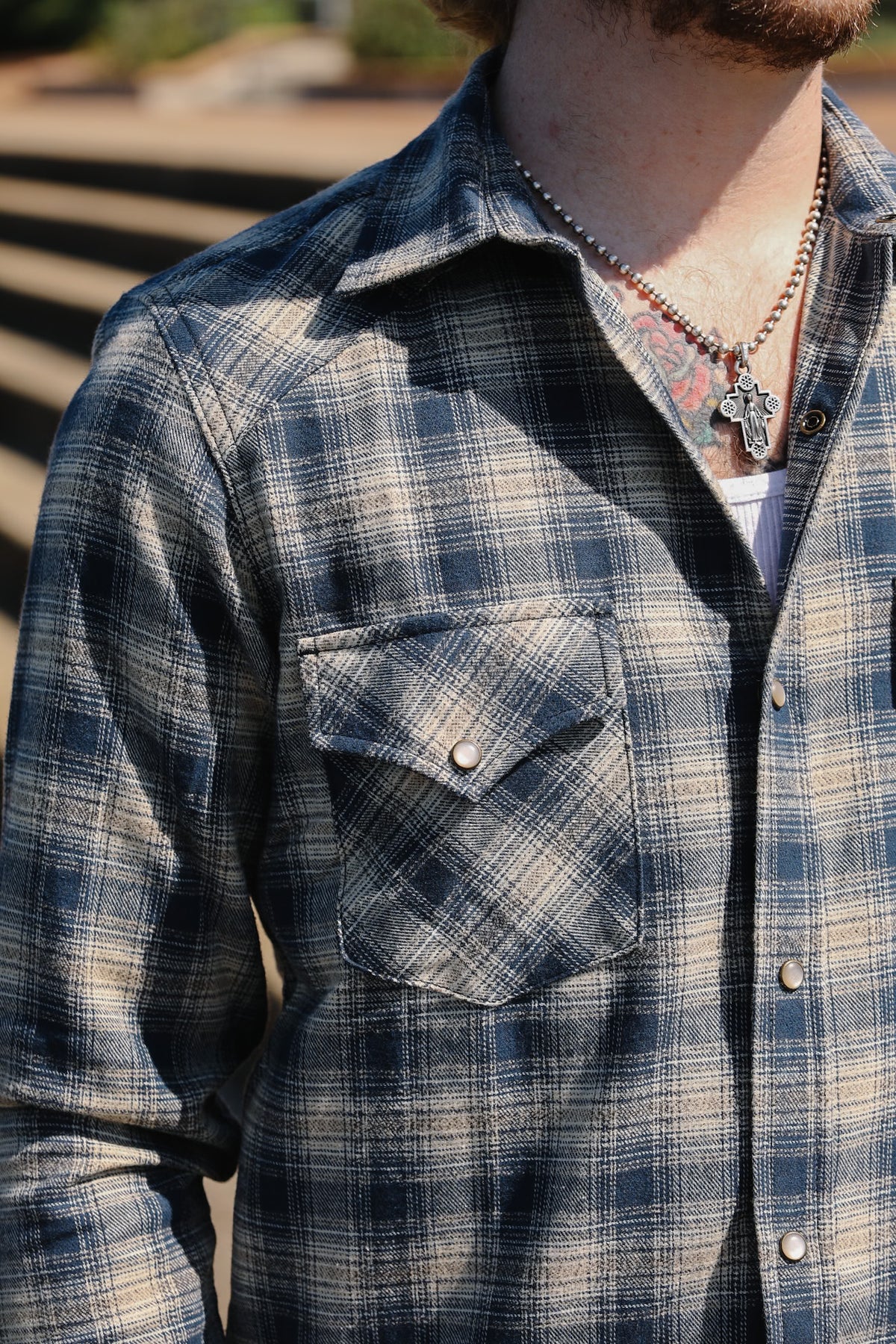 A close-up of a man wearing a blue and beige plaid button-down flannel shirt, styled with a beaded necklace and a silver pendant.