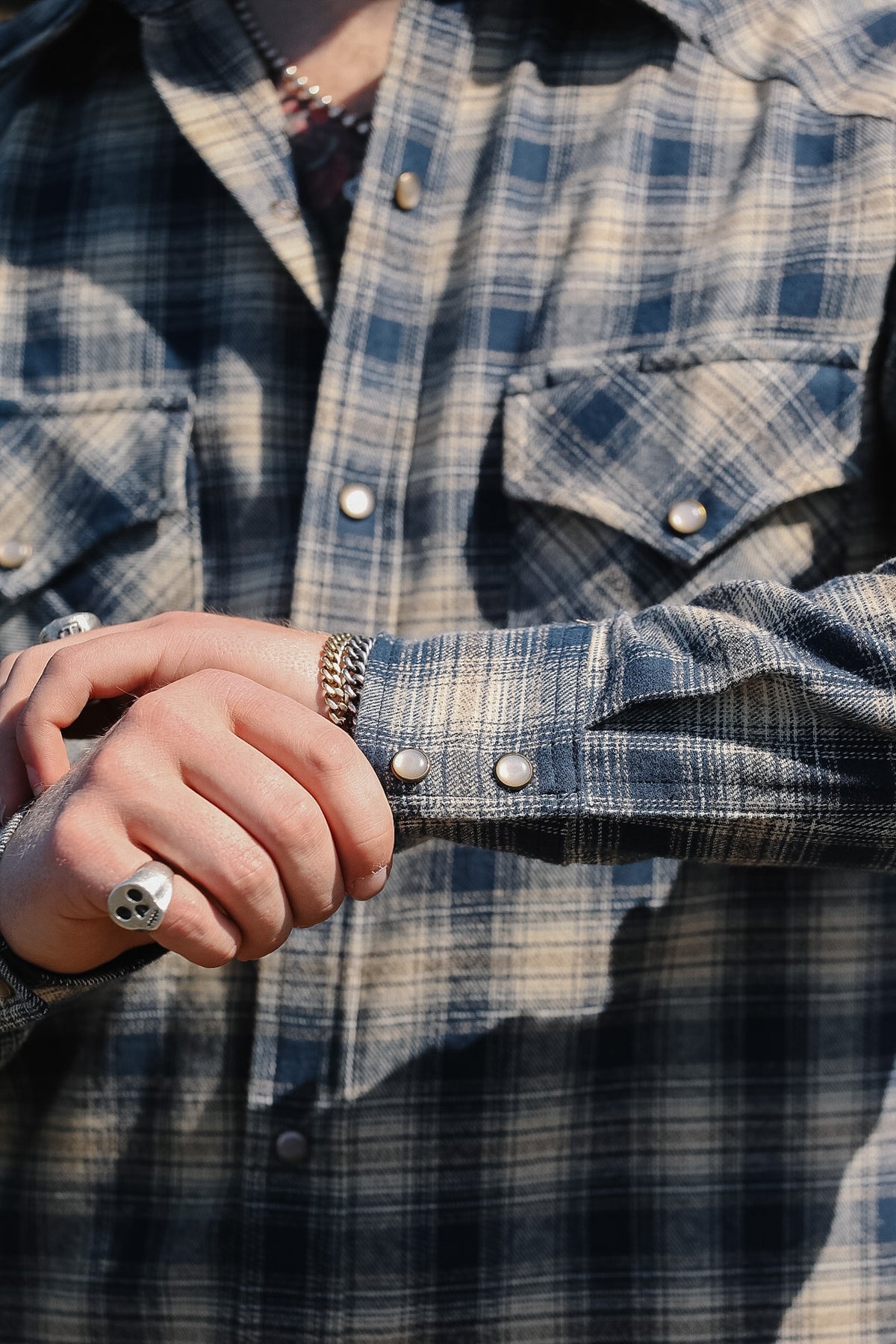 Close-up of a person wearing a blue and beige plaid flannel shirt, adjusting the cuff with one hand while wearing silver rings.