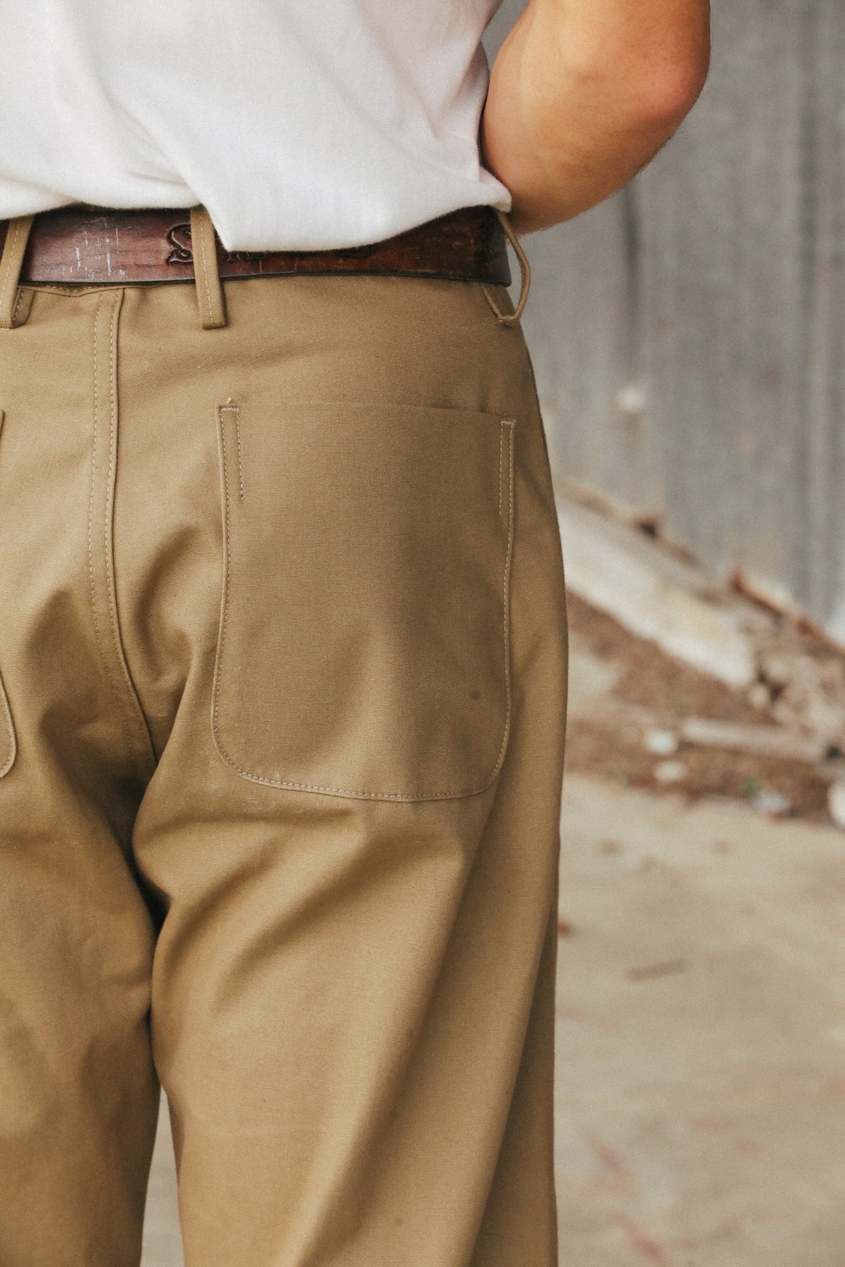 Close-up of a person wearing tan work pants with a brown belt and white shirt, standing against a blurred outdoor concrete background.