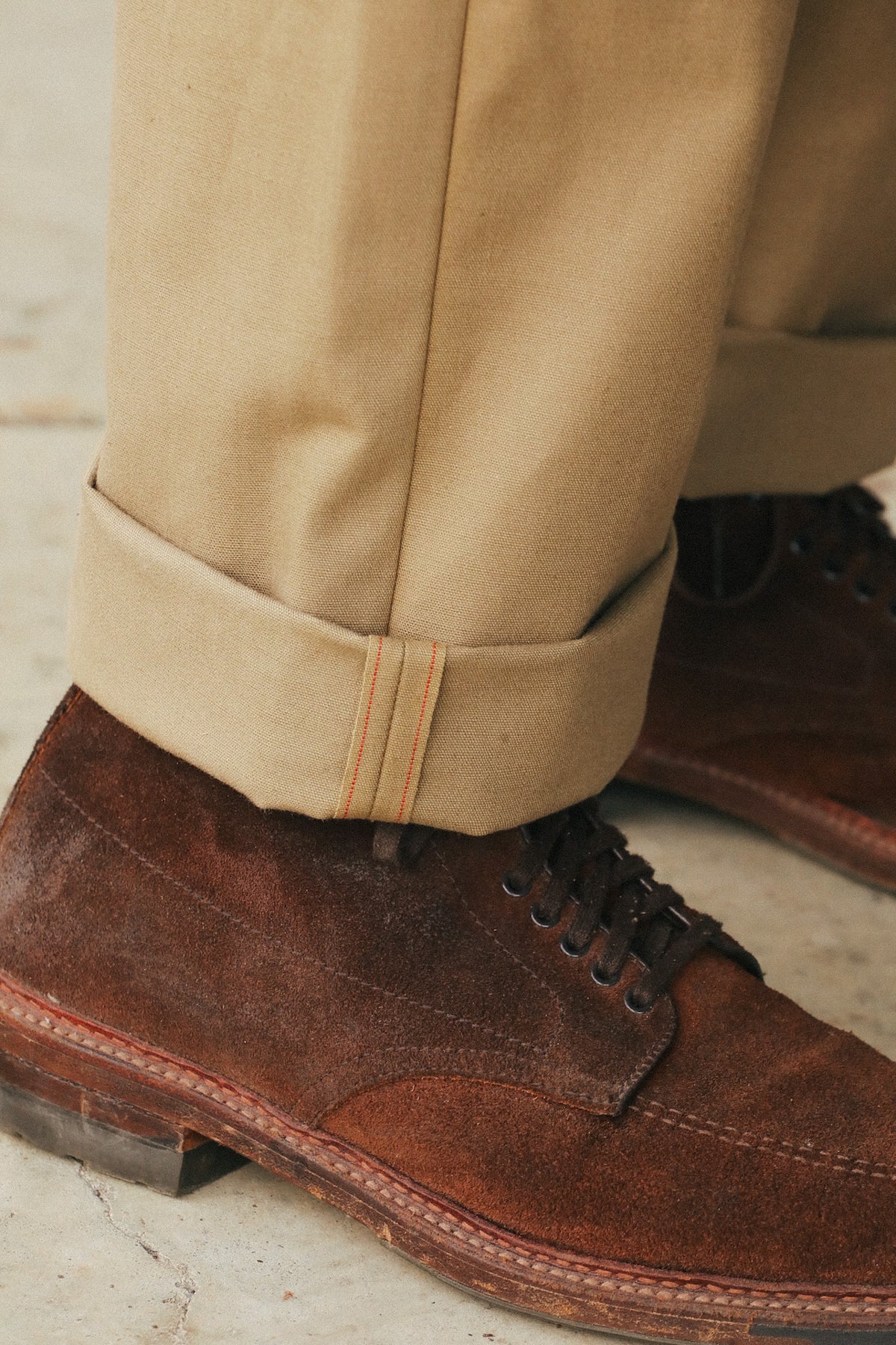 A close-up of a person's legs wearing tan cuffed trousers and dark brown suede lace-up shoes on a light stone floor.