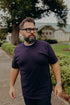 Bearded man wearing black glasses and a dark purple t-shirt outdoors with residential houses and greenery in the background