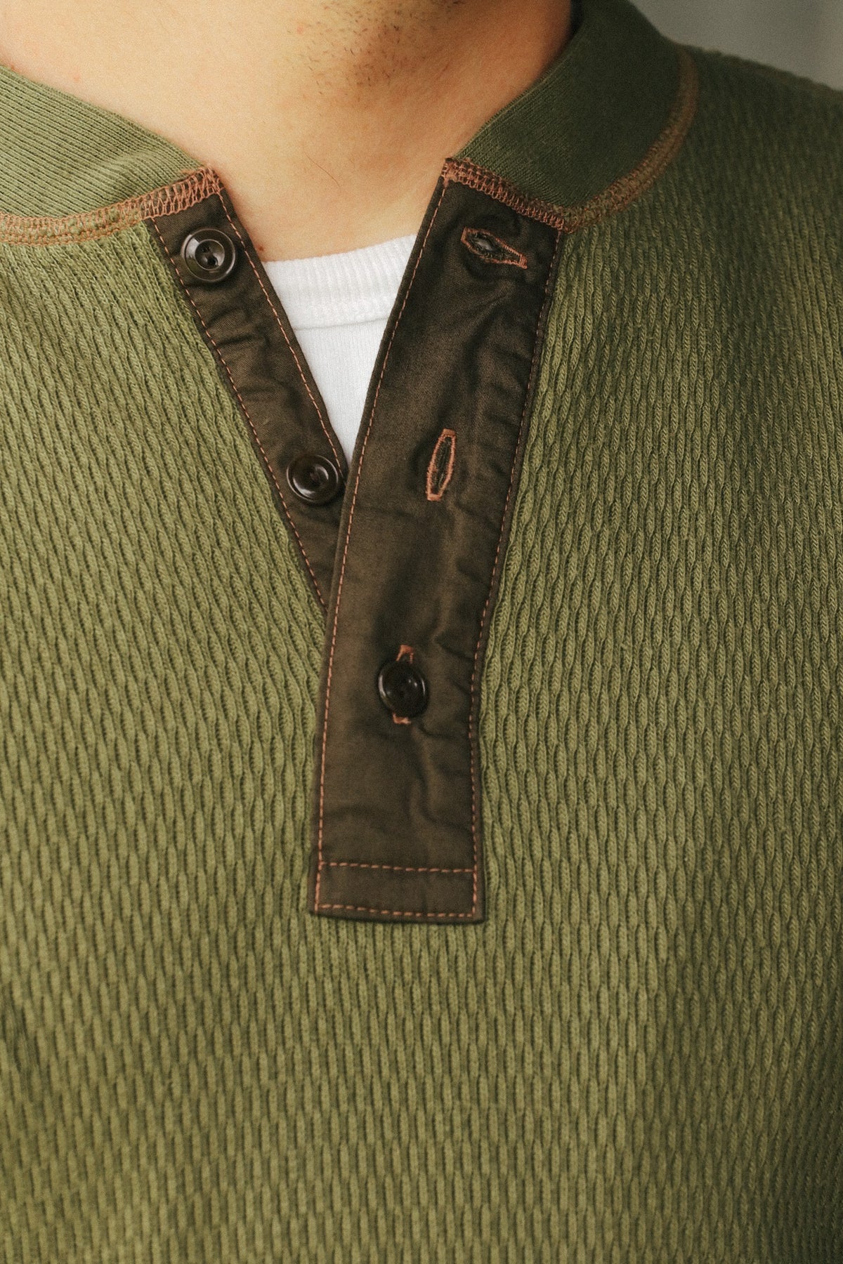 Close-up of a textured olive green henley shirt with a contrasting dark brown button placket and a white undershirt visible at the collar.