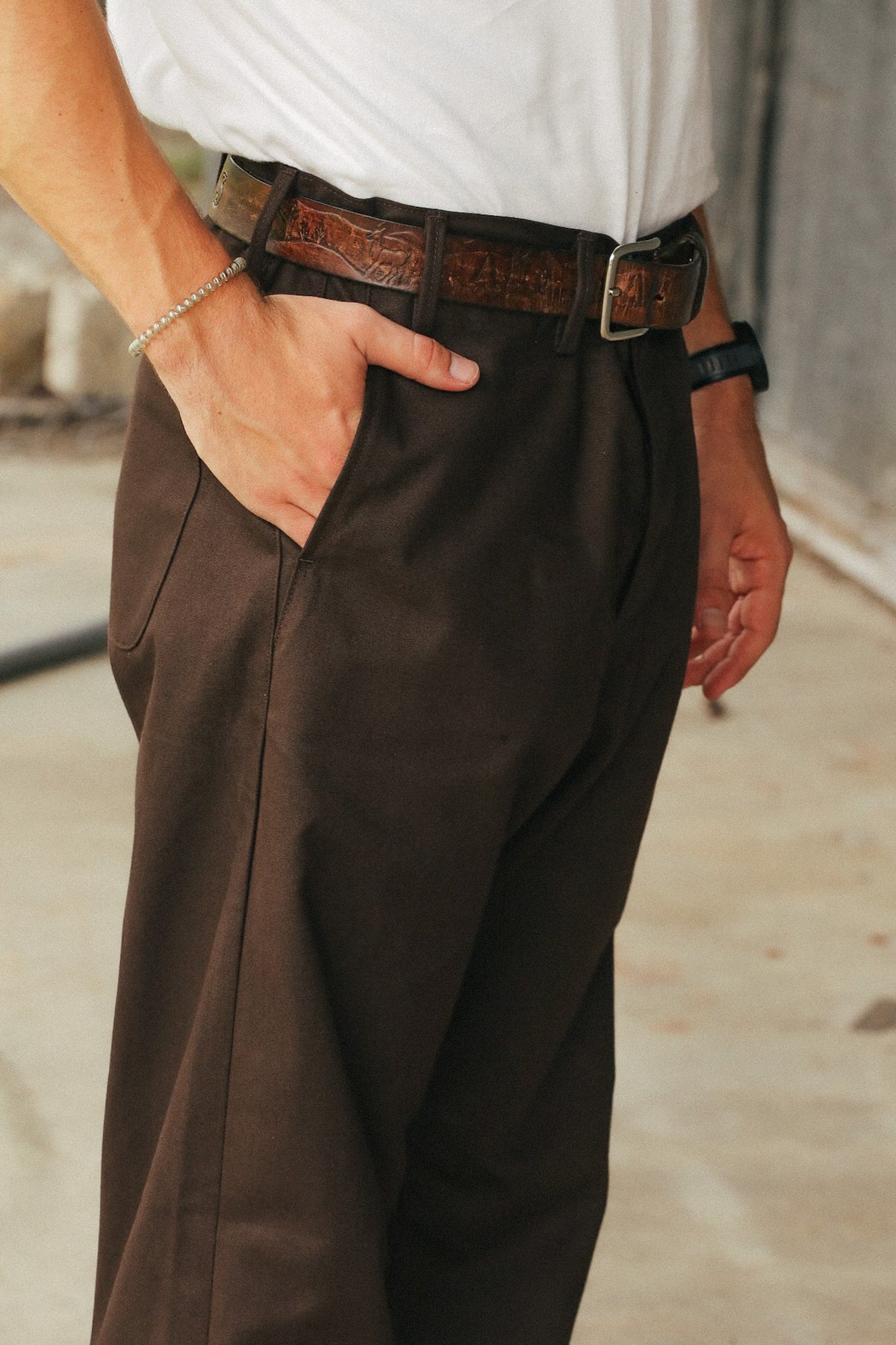 A close-up of a person wearing dark brown dress pants with a brown embossed leather belt, a white shirt, and accessories, standing in an outdoor concrete setting.