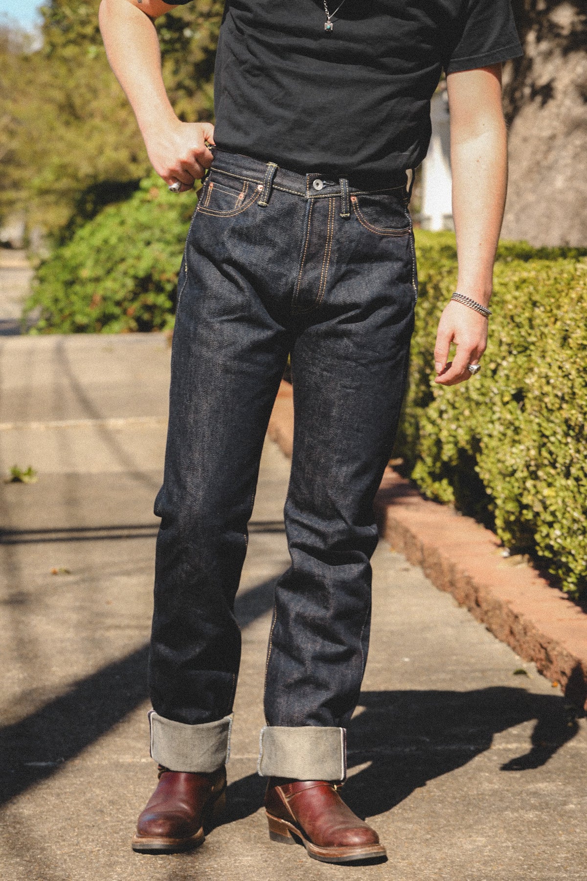 A person outdoors wearing dark indigo denim jeans with rolled cuffs, a black t-shirt, and brown leather boots on a paved walkway with green hedges in the background.