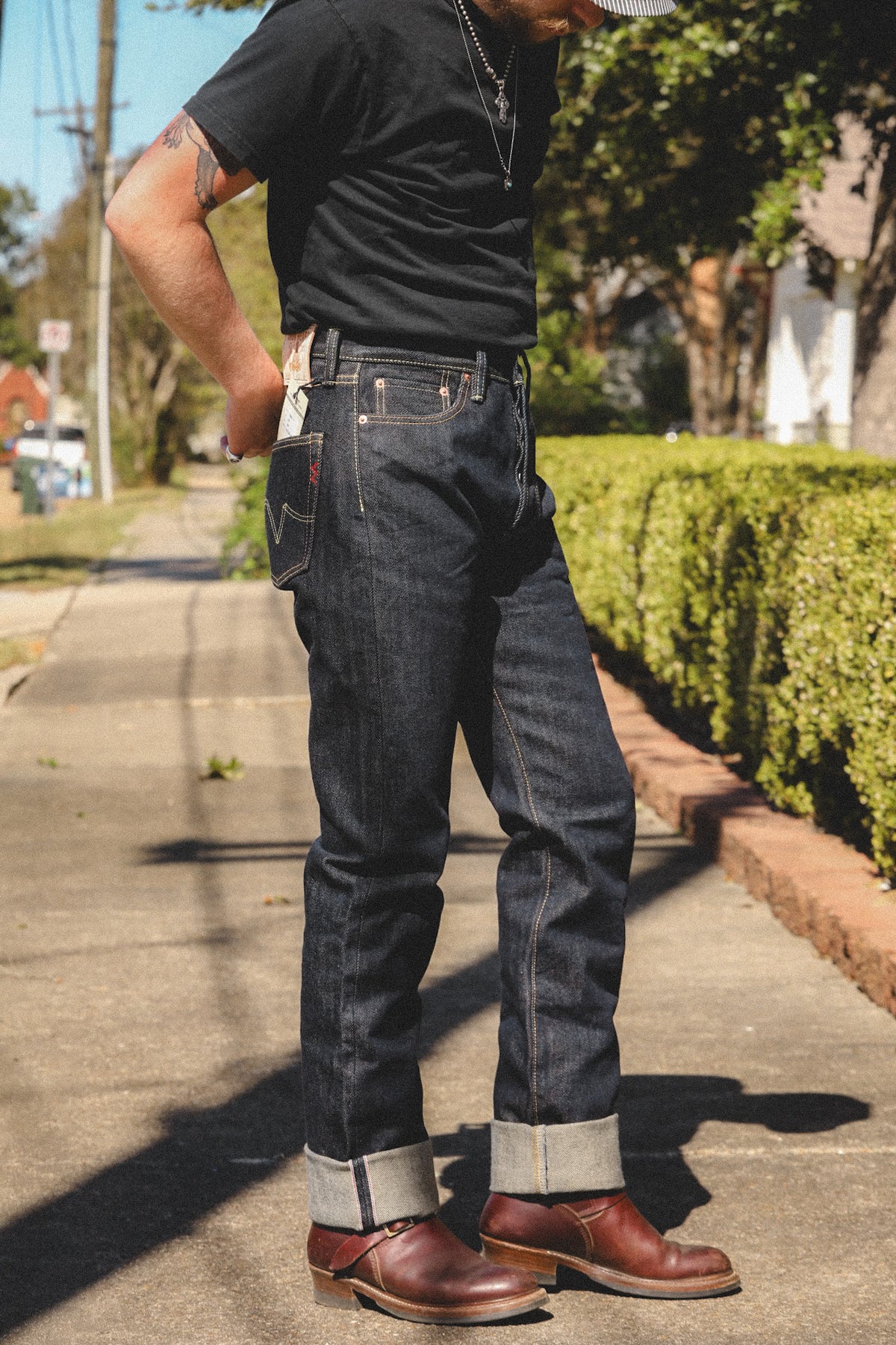 A person standing outdoors on a sidewalk wearing dark indigo denim jeans with rolled cuffs, a black t-shirt, and brown leather boots. The background shows a suburban street with green hedges and trees.