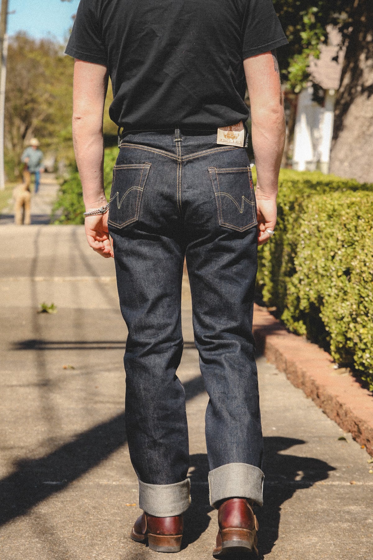 A person viewed from behind wearing dark indigo denim jeans with rolled cuffs, a black t-shirt, and brown leather boots, standing on an outdoor paved path.