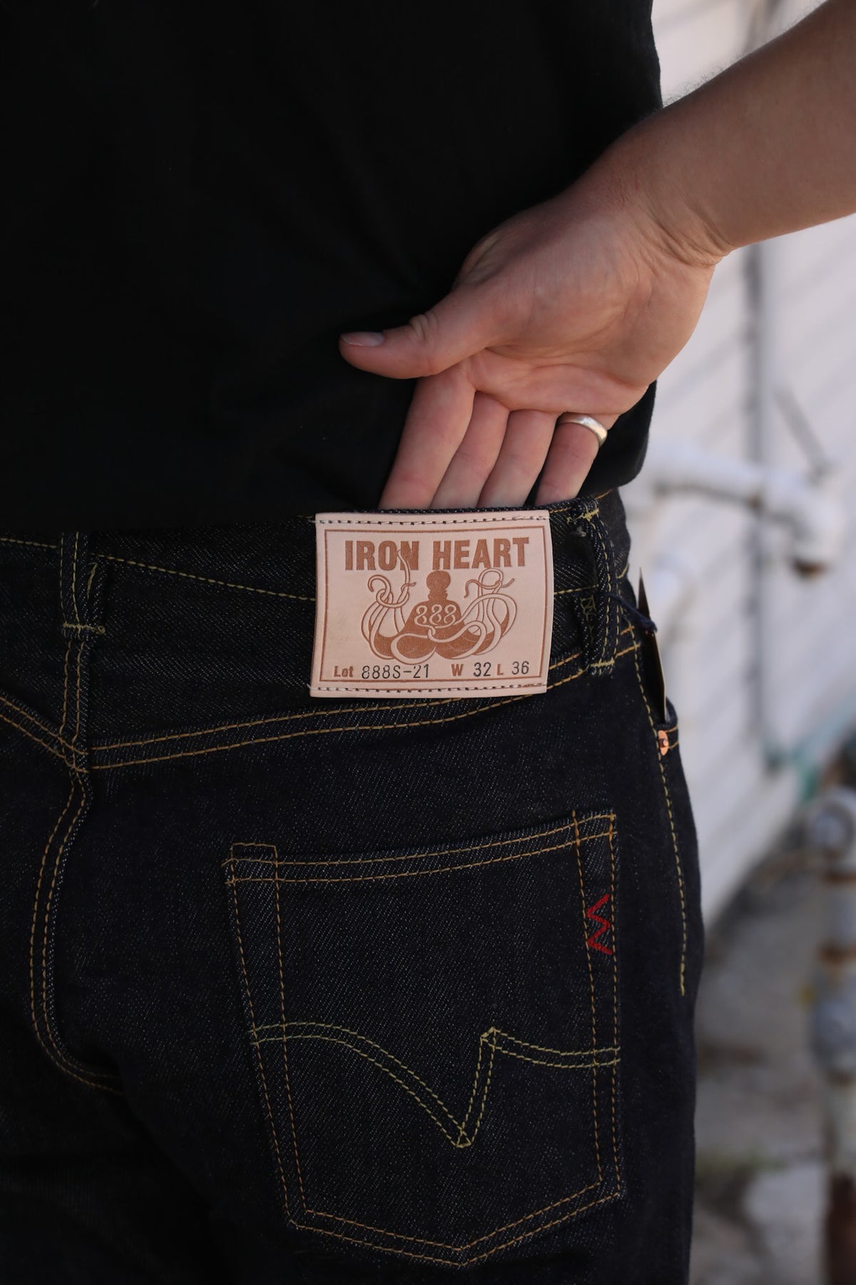 Close-up of a person's hand pulling the waistband of dark indigo jeans to reveal a beige leather patch with the brand name 'IRON HEART'.