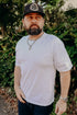Man wearing white t-shirt, black cap with circular logo, silver chain necklace, and watch standing outdoors with green foliage background