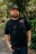 Man wearing black cap with floral logo, black t-shirt, and wristwatch standing outdoors near green foliage