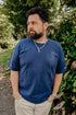 Bearded man wearing a blue T-shirt and silver chain necklace standing outdoors near greenery
