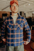 Man wearing blue plaid flannel shirt, beige t-shirt, red cap, and green pants indoors at Iron Shop Provisions