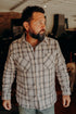 Bearded man wearing a brown and beige plaid button-up shirt indoors with a dog in the background