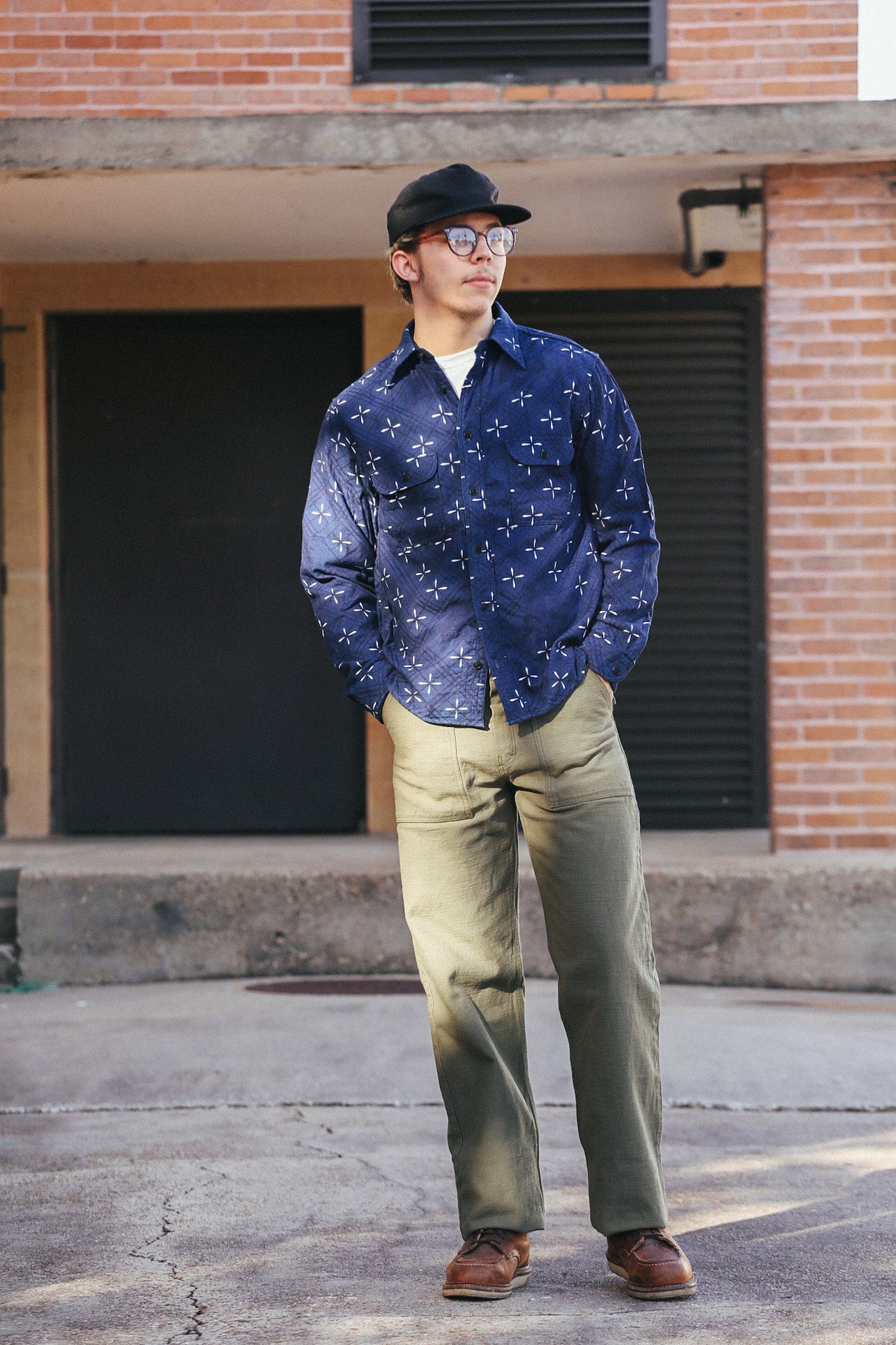 Man wearing a blue patterned shirt and green pants standing in front of a brick building.